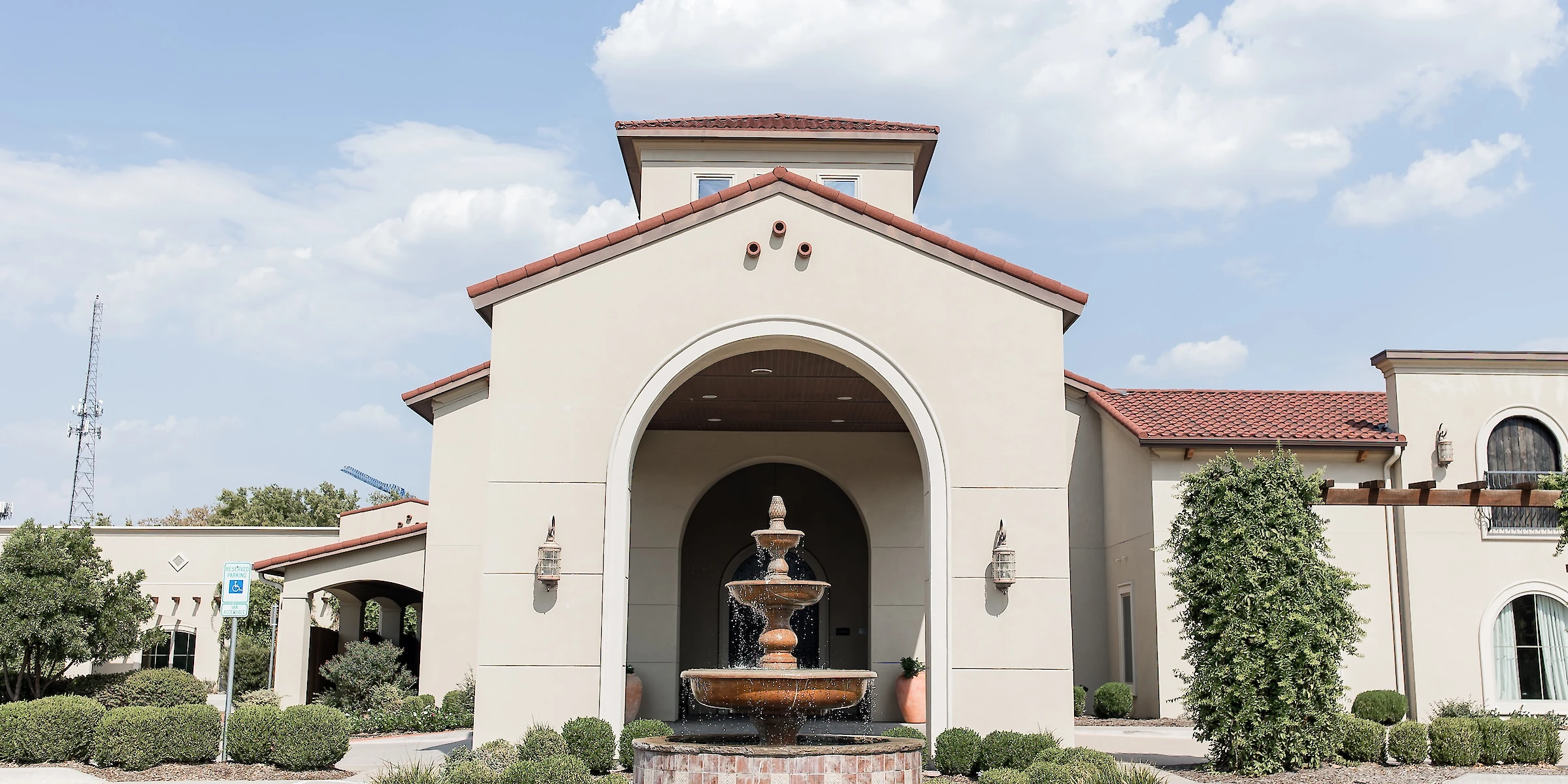 Exterior entrance of a Mediterranean-style event venue with tan stucco walls and a terracotta tile roof, featuring a central arched entryway and a large tiered copper water fountain.