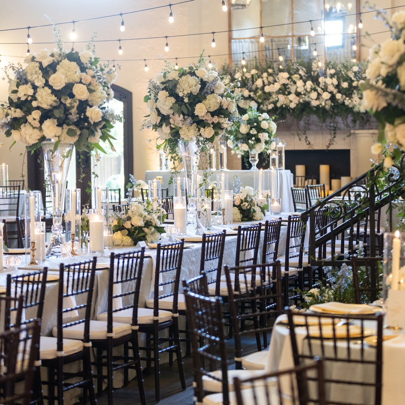 A luxurious wedding reception room with long tables set with tall white and light blue floral arrangements, candles, and string lights overhead.