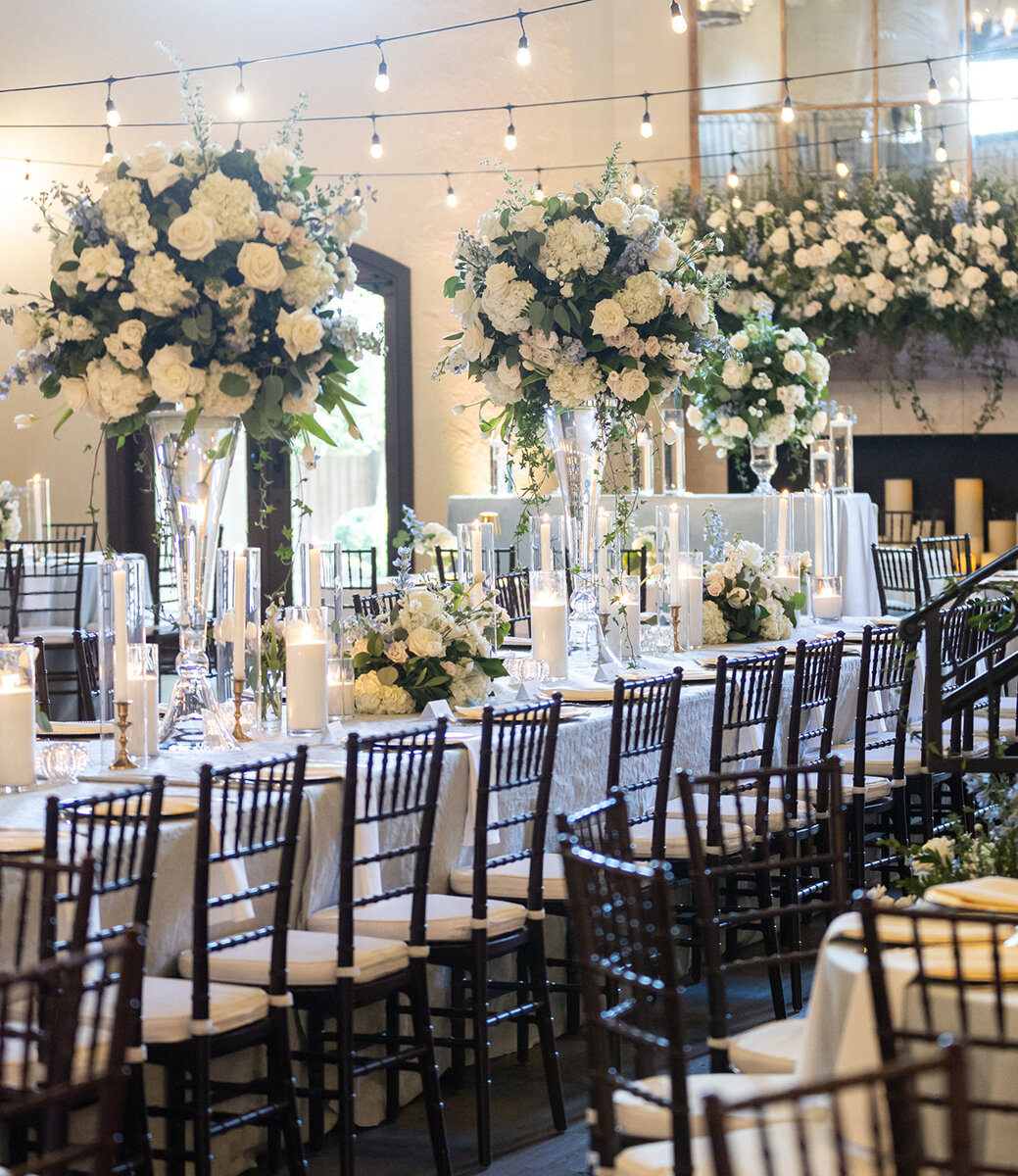 Elegant indoor wedding reception decorated with tall white floral centerpieces, candlelit tables, string lights, and dark wood chairs