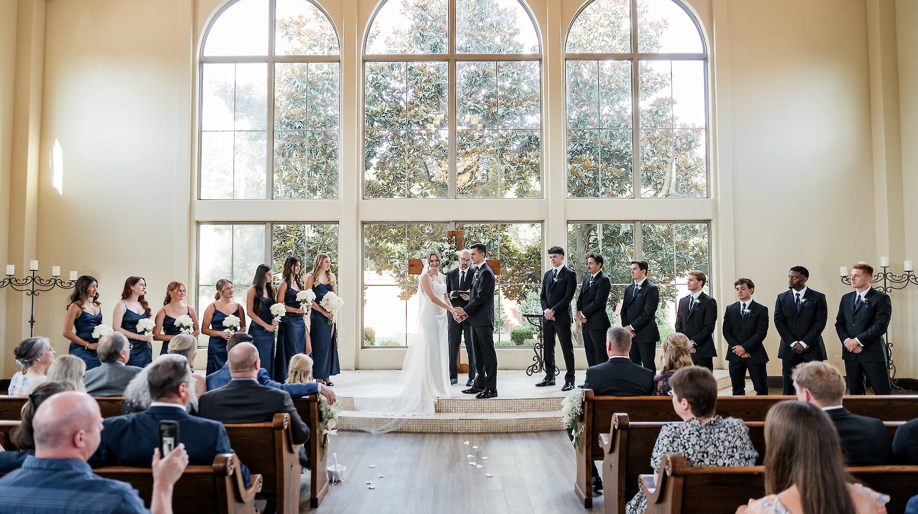 Wide view of a wedding ceremony taking place in a modern chapel with a vaulted ceiling and a dramatic wall of three tall arched windows overlooking trees. The wedding party stands on a raised altar, with bridesmaids in navy blue dresses and groomsmen in black tuxedos.