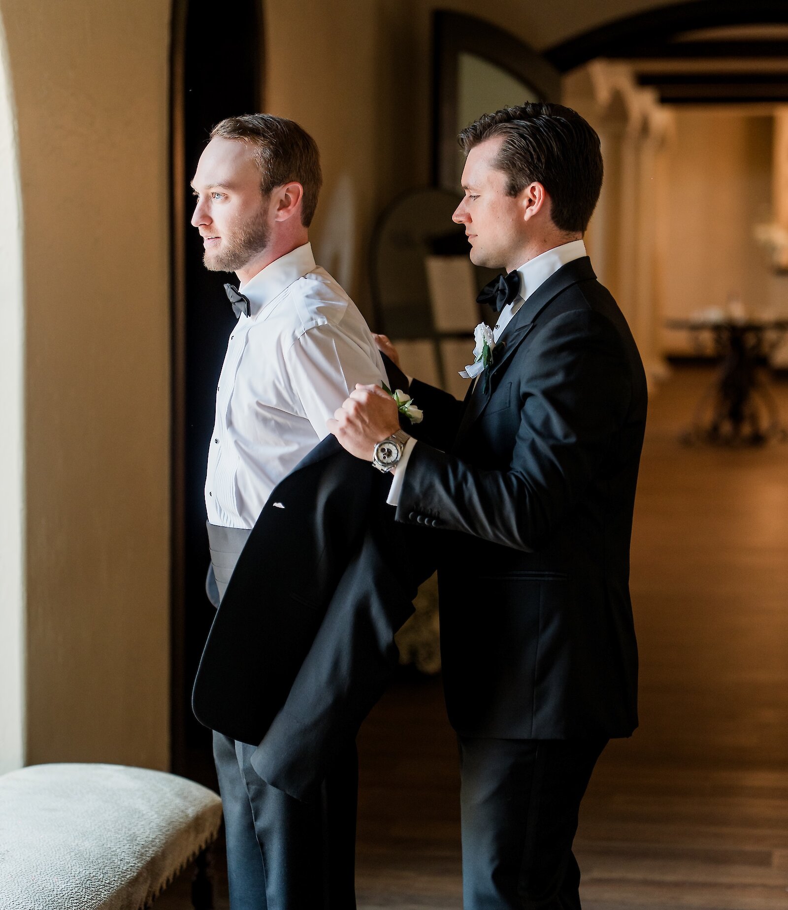 Groomsman helping the groom put on his black tuxedo jacket and white shirt in a dimly lit, arched hallway. The groom is wearing a black bowtie and a white boutonnière.