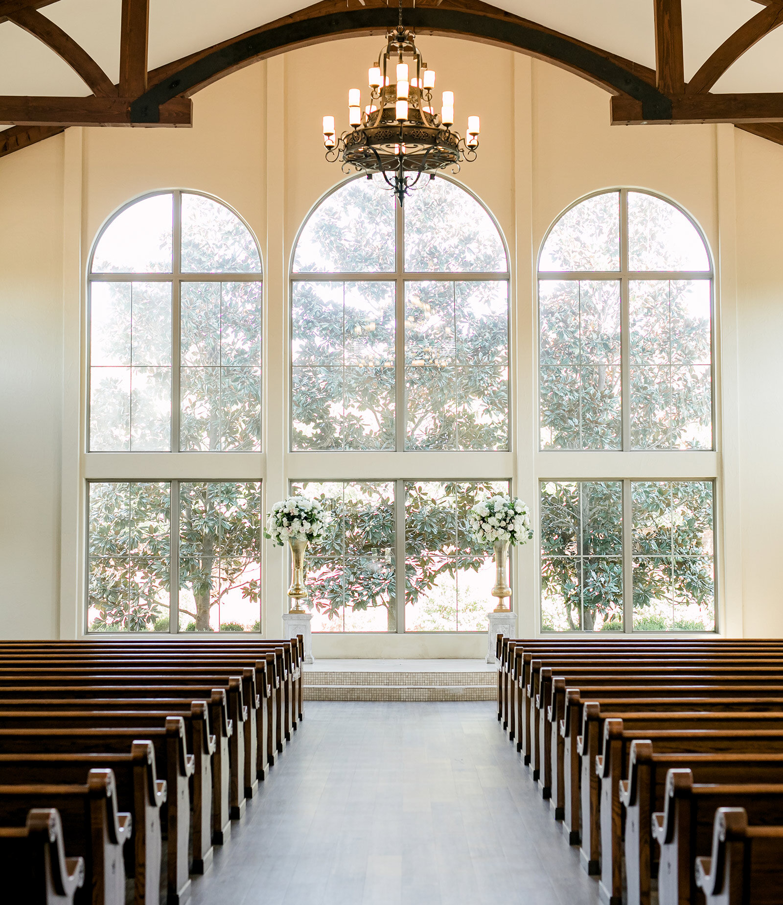 Elegant wedding chapel interior with a vaulted white ceiling featuring exposed dark wood trusses. The aisle is lined with wooden pews and large, lush floral arrangements featuring peach, coral, and white roses, set in front of a dramatic wall of arched windows.