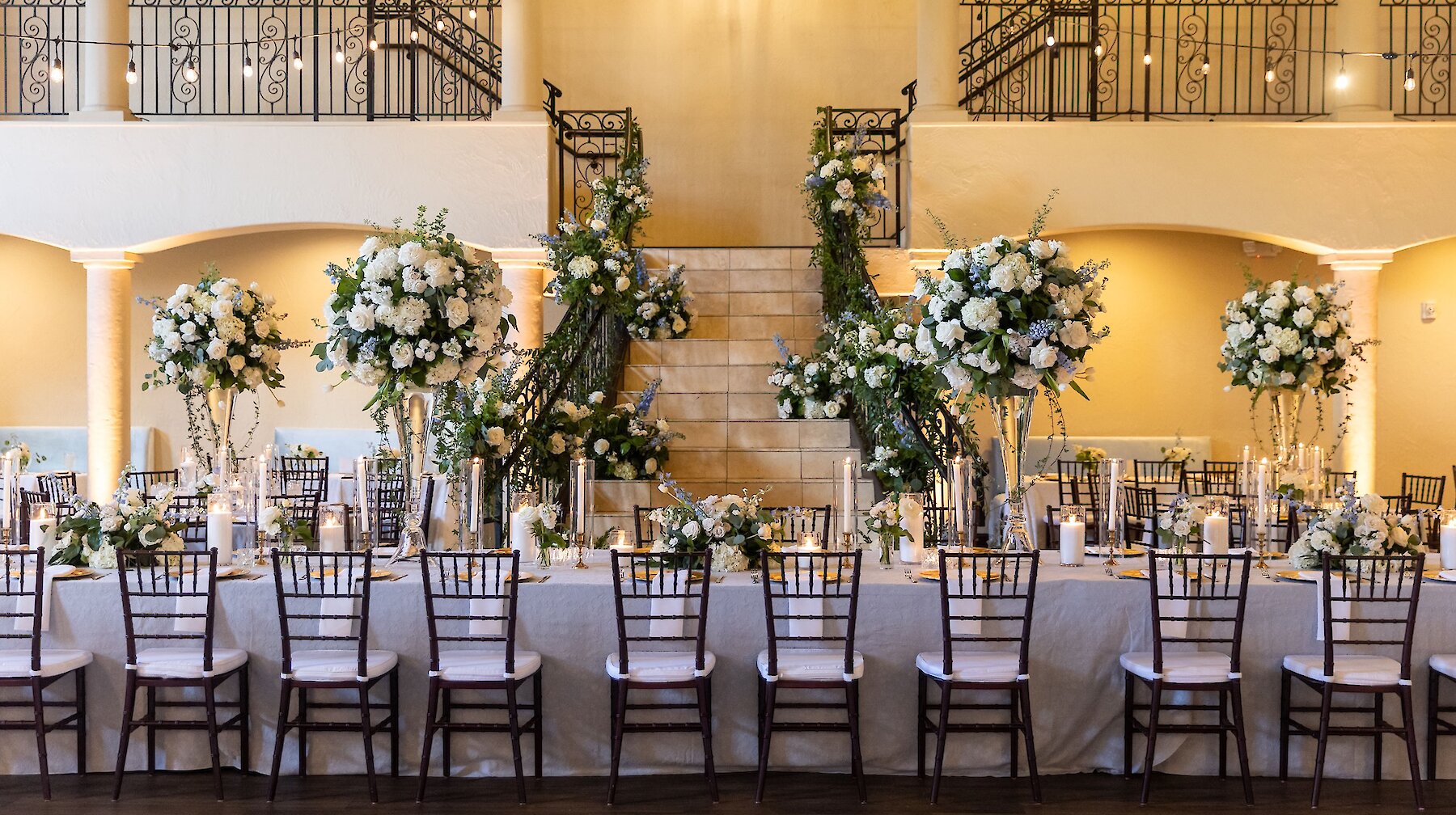 n elegant wedding reception hall with a long head table, chiavari chairs, tall white and green floral centerpieces, and a grand staircase decorated with flowers.
