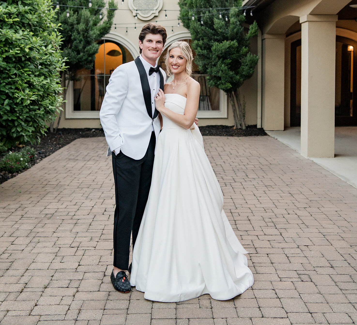 Stylish wedding portrait of the couple posing on a brick paver courtyard under string lights. The groom wears a white tuxedo jacket and black trousers, and the bride wears a classic strapless white satin ballgown.