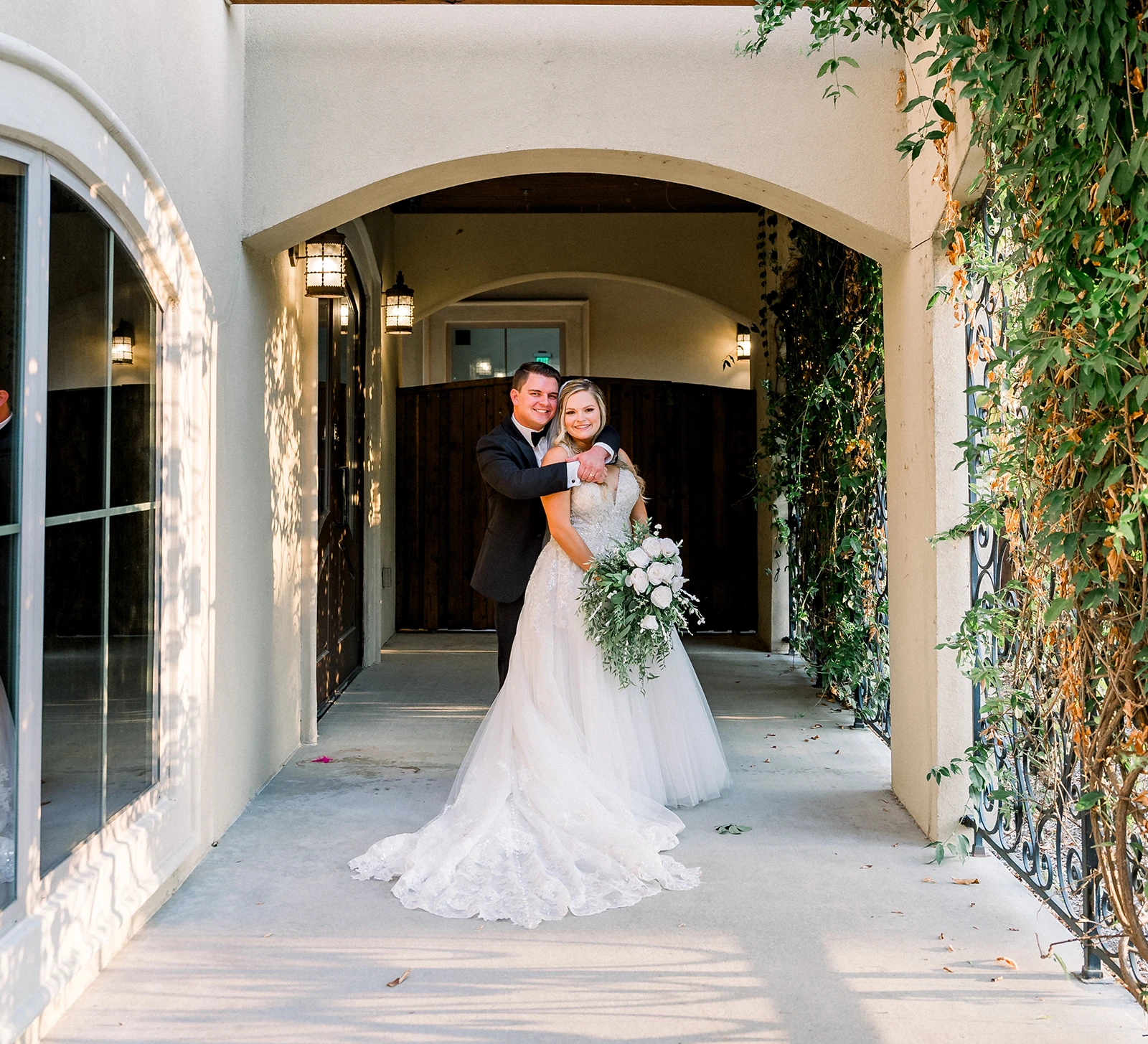 Bride and groom posing under an arched outdoor walkway with greenery on the sides, the bride holding a white floral bouquet