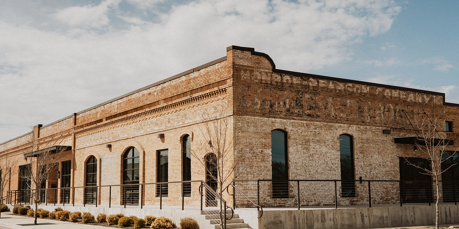 Exterior of a historic industrial building or loft venue featuring light orange brick, arched windows, and faint painted vintage signage reading "MILLER BROACH COMPANY" near the roofline, under a bright, cloudy sky.