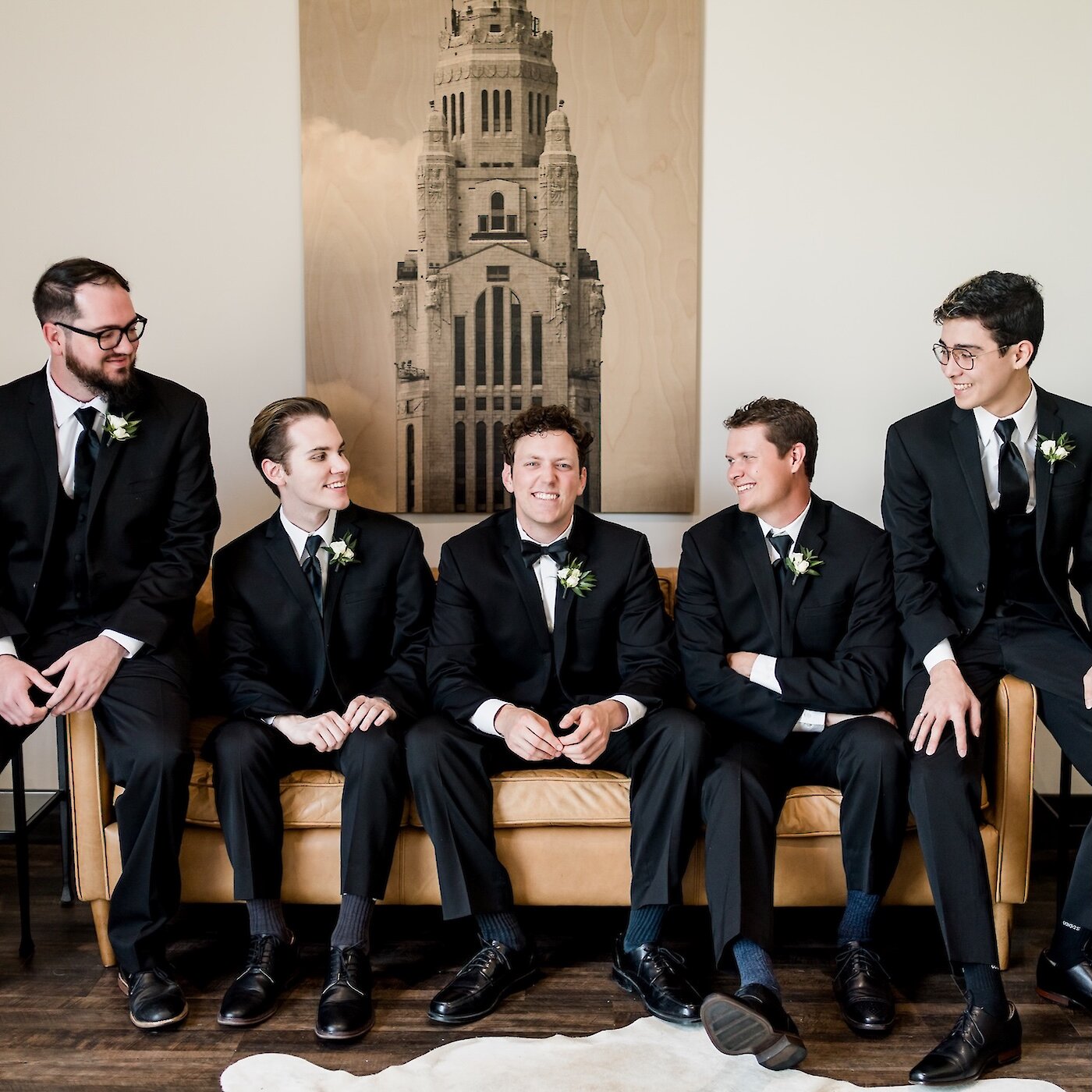 Group portrait of the groom and four groomsmen smiling while seated on a cognac leather couch. All men wear black tuxedos and white boutonnieres in a modern lounge area with a large architectural artwork on the wall.
