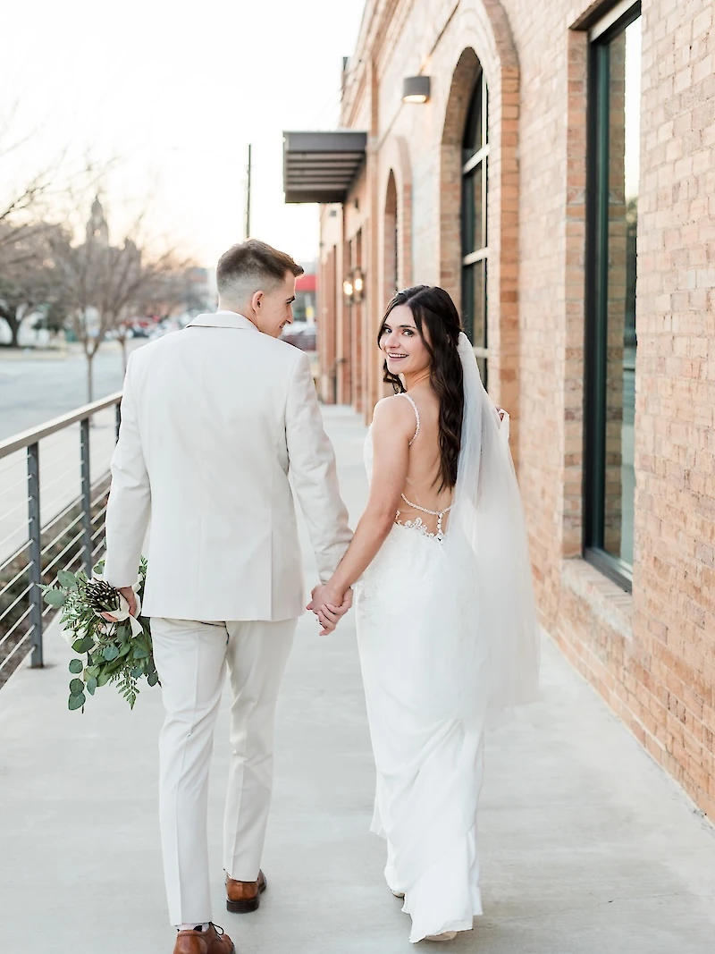 Sunlit wedding portrait of the couple walking away down a brick-lined sidewalk next to a rustic brick building with arched windows. The groom wears a tan suit, and the bride smiles back, showing her low-back lace gown and simple veil.