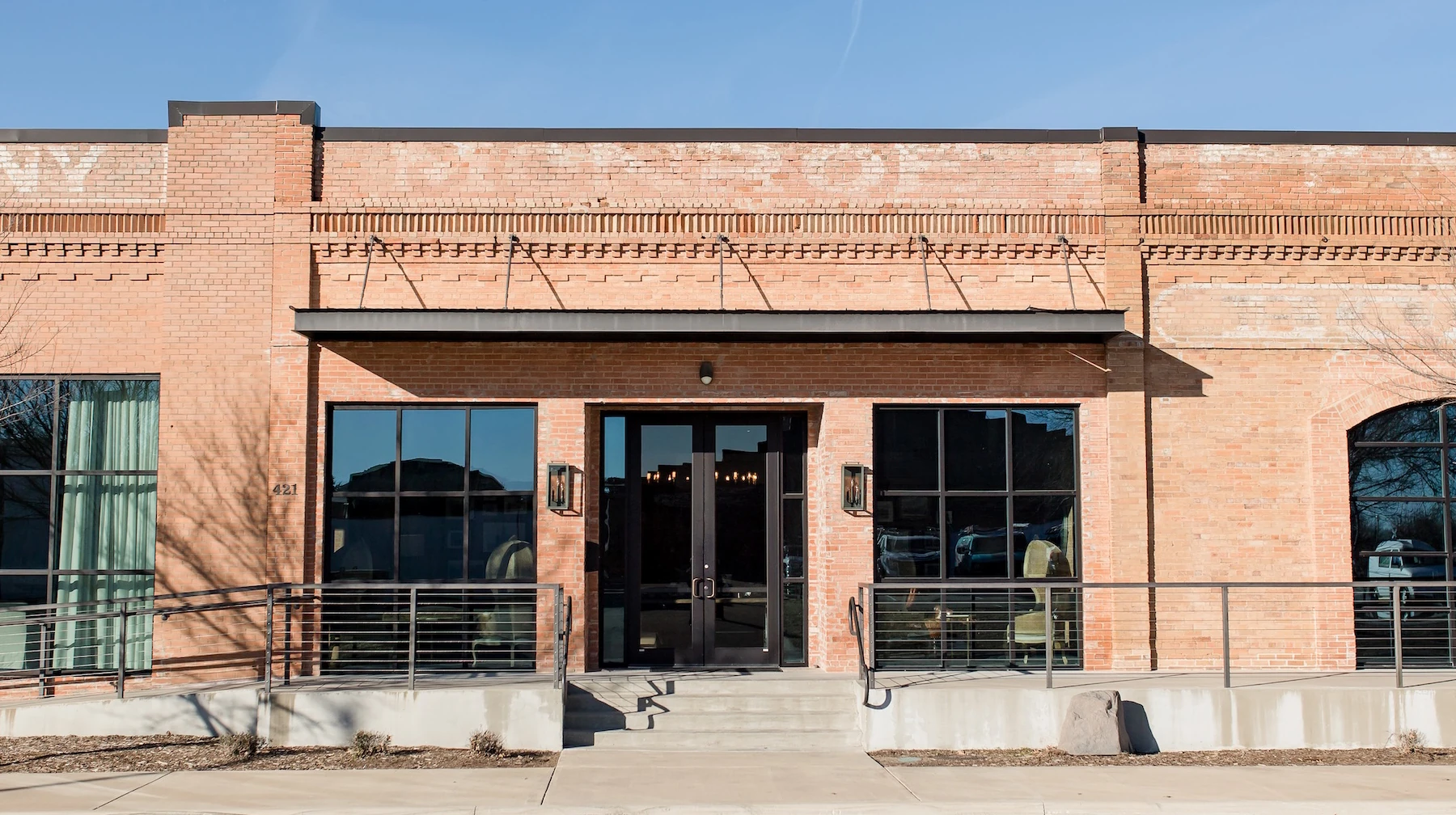 Exterior of an industrial brick building with large black-framed windows, double glass doors, and metal railing, shown on a clear sunny day
