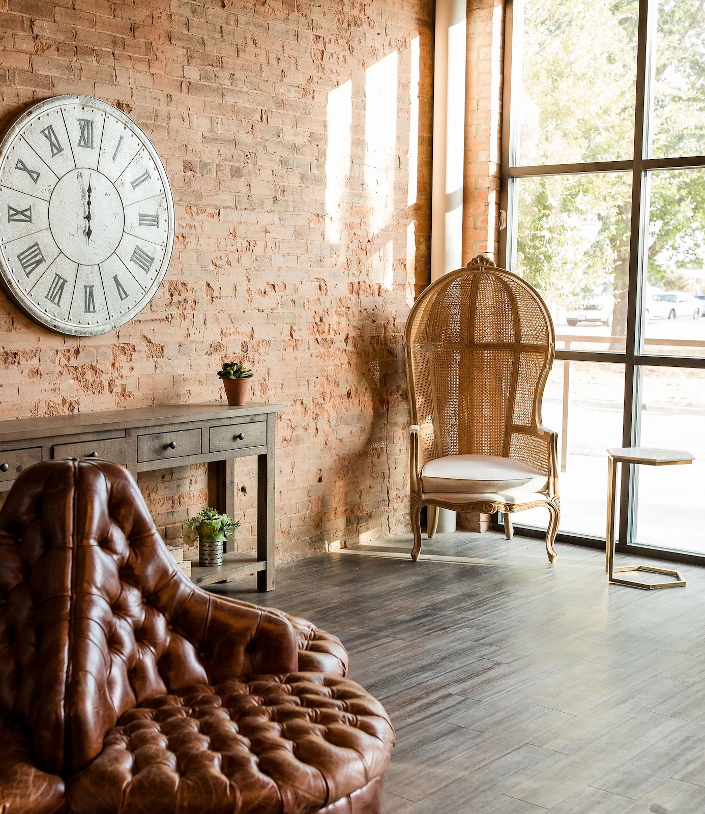 Industrial-style lounge with exposed brick wall, large Roman numeral clock, leather tufted seating, and a wicker accent chair beside floor-to-ceiling windows
