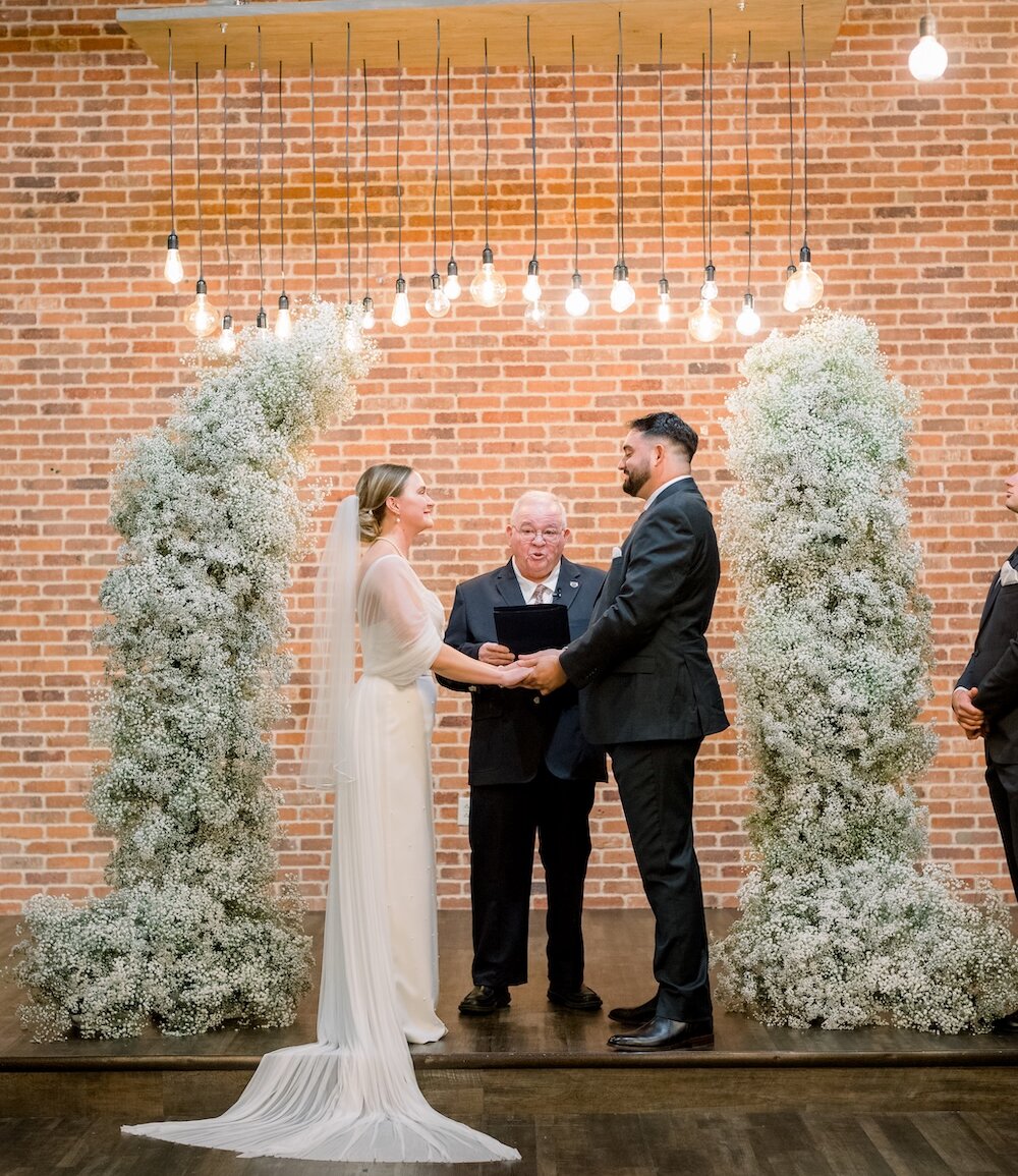 Intimate wedding ceremony at an altar with an exposed brick wall backdrop. The couple stands between two tall, voluminous baby's breath floral pillars under a canopy of Edison string lights, exchanging vows with the officiant.