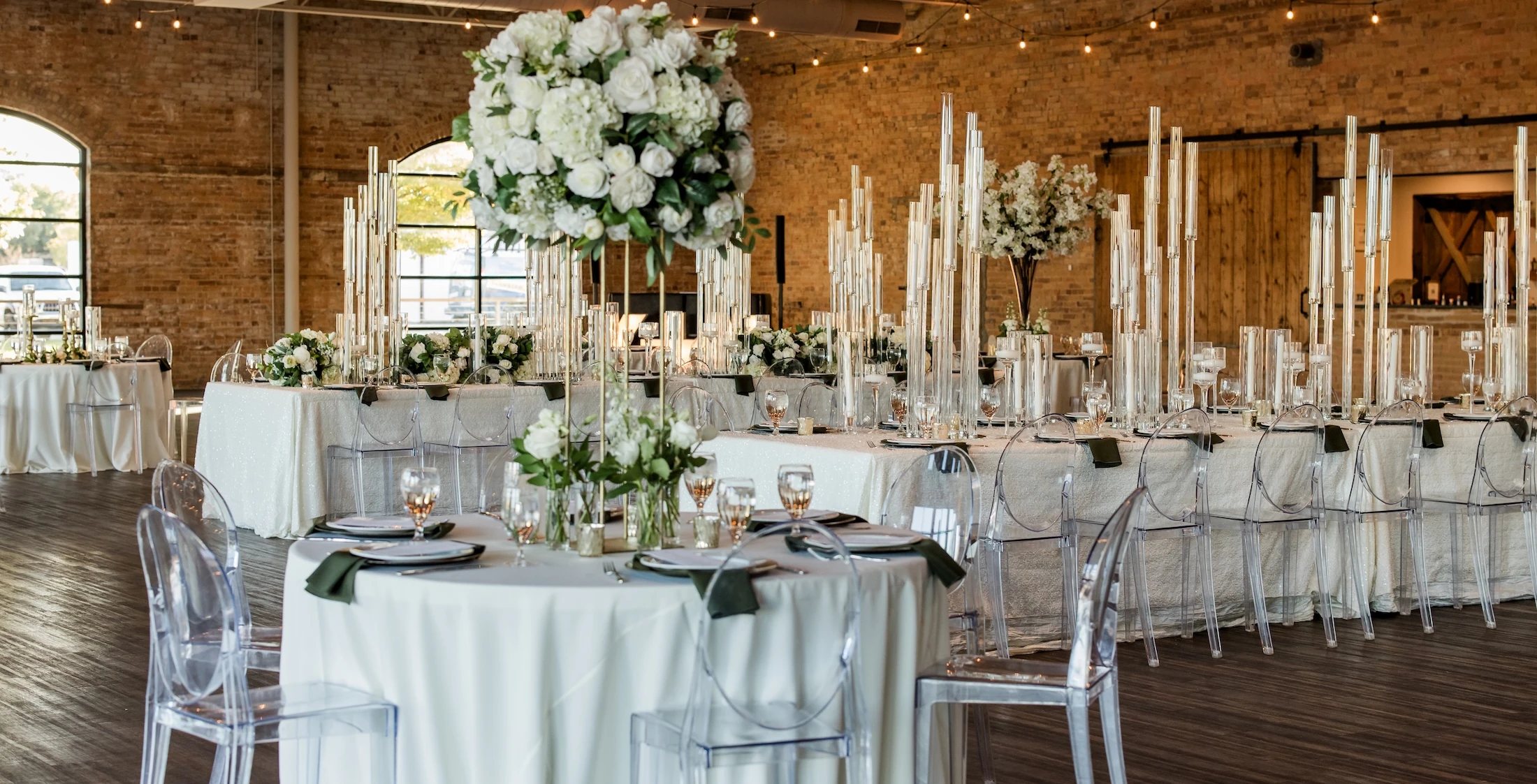 Elegant wedding reception setup in a modern industrial venue with tall white floral centerpieces, clear acrylic chairs, candle arrangements, and soft neutral décor.