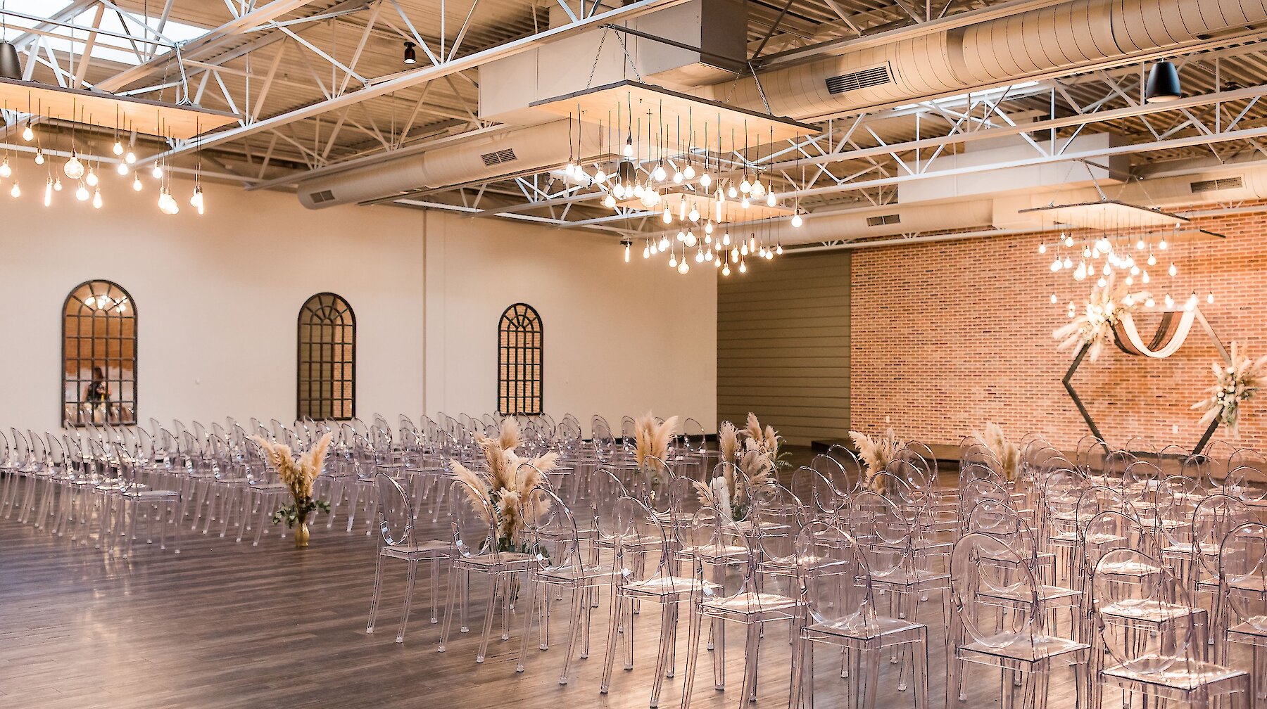 An industrial-style wedding ceremony space with exposed ceiling trusses, acrylic ghost chairs, pampas grass decor, and arched windows.
