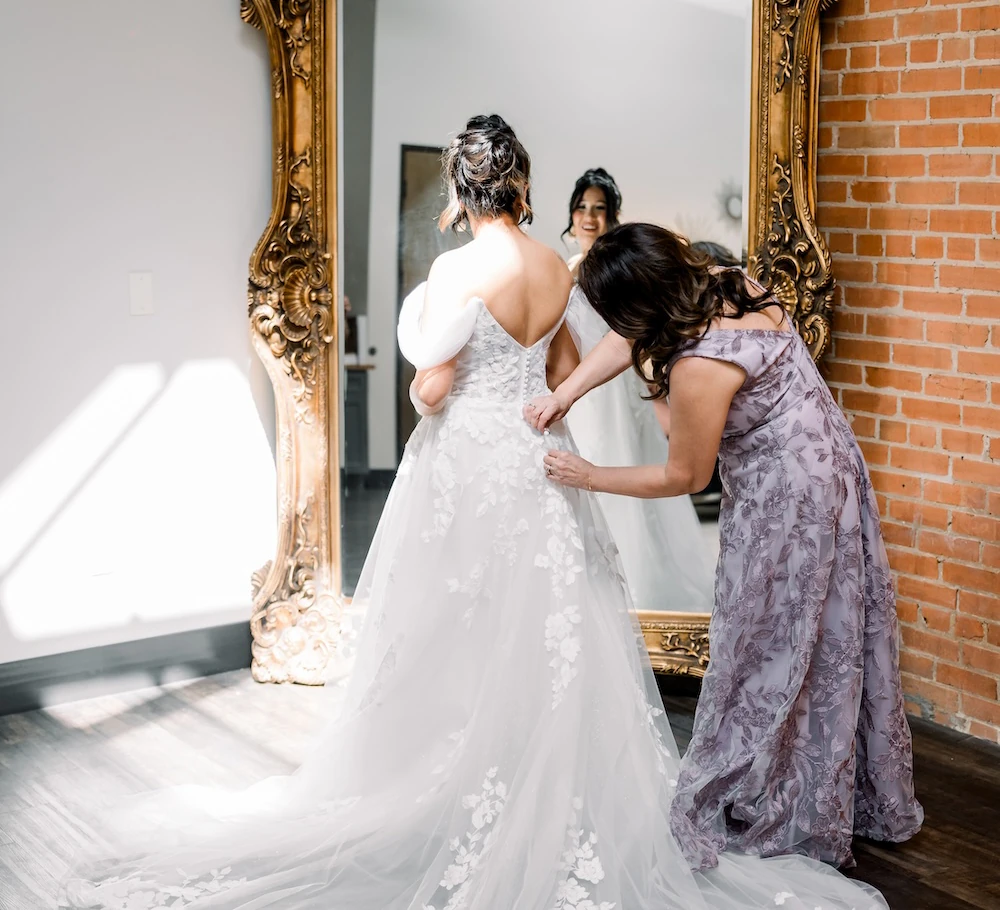 The bride, wearing a white floral lace gown, is being helped into her dress by a woman in a purple dress in front of a large, ornate gold mirror.