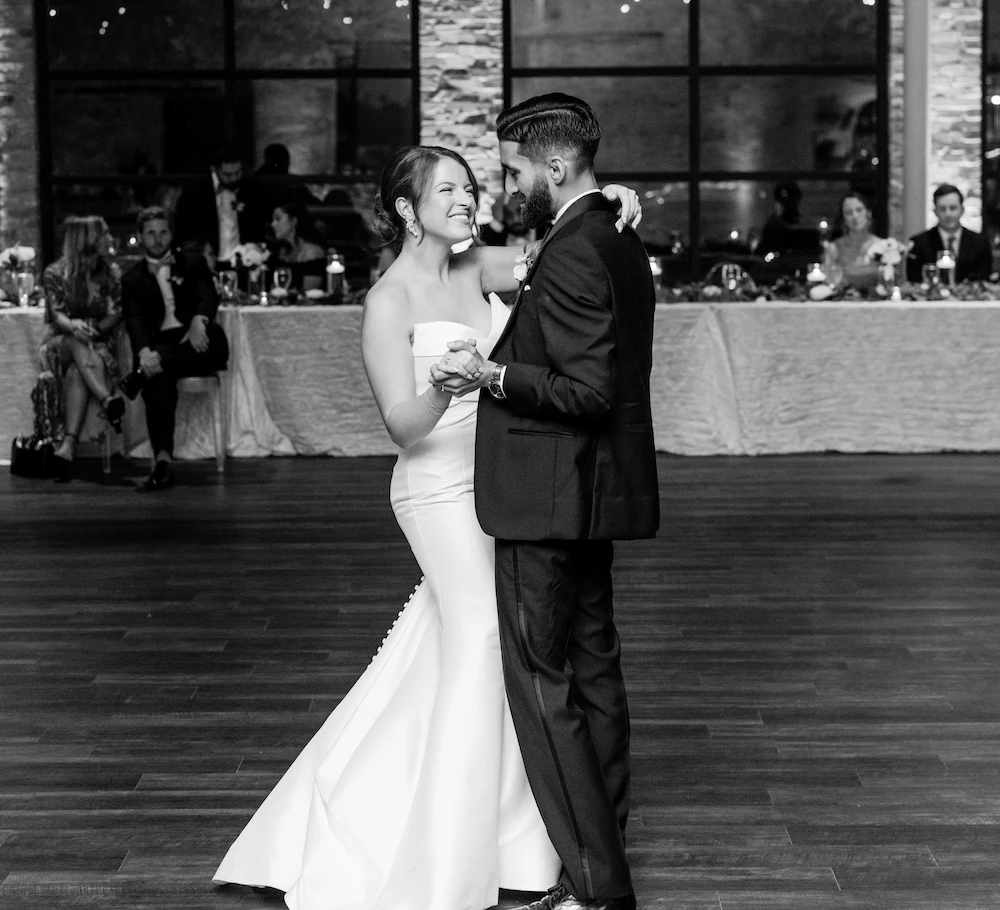 Bride and groom sharing their first dance in an elegant indoor wedding venue, smiling warmly at each other on a wooden dance floor with guests seated in the background