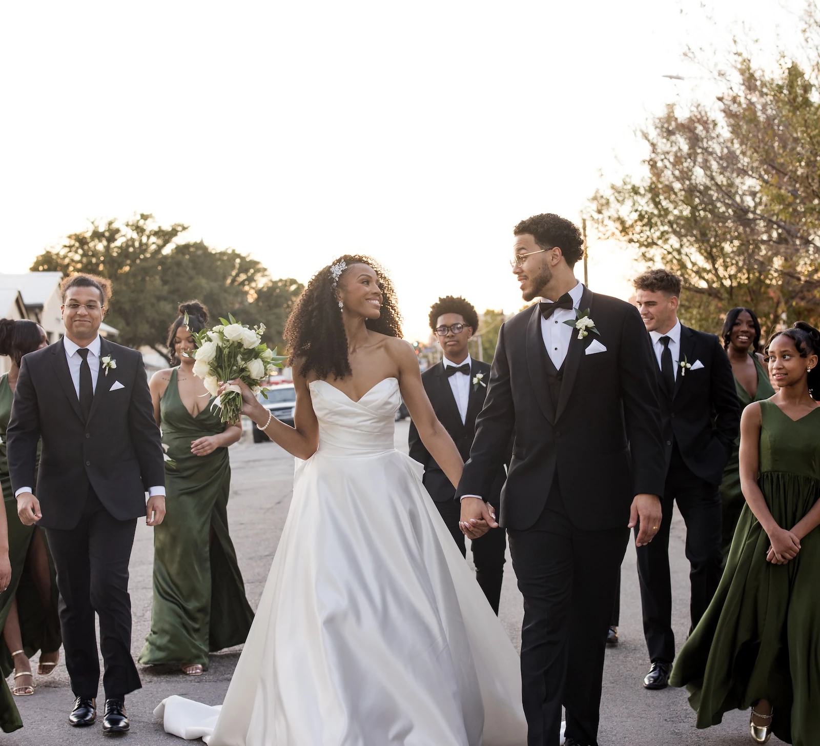 Joyful portrait of the bride and groom leading their wedding party down an urban street at sunset. The groom wears a black tuxedo, and the bridesmaids wear various styles of dark olive green satin gowns, walking alongside groomsmen in matching black tuxedos.