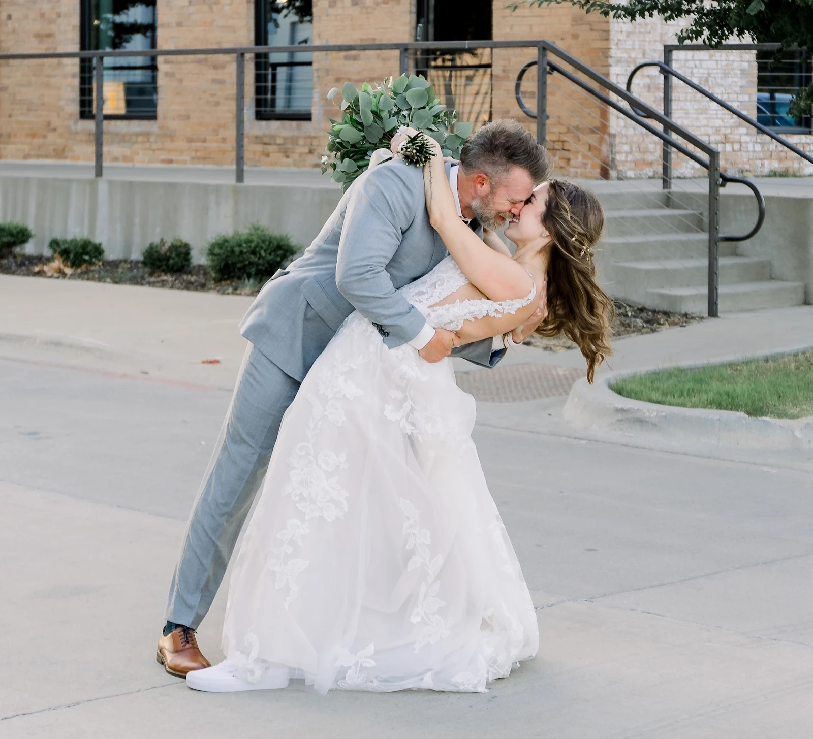 Groom dipping and kissing bride outdoors in front of a rustic brick building, with the bride holding a bouquet and her lace wedding gown flowing