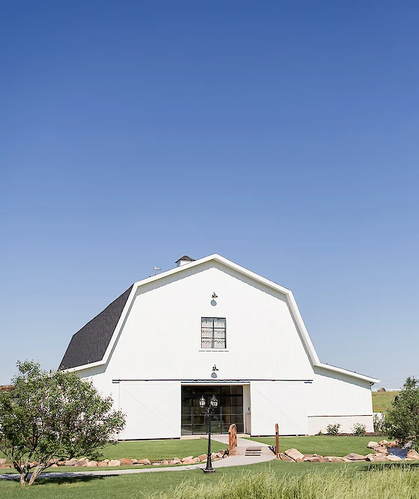 White barn-style building with dark roof shingles, large sliding doors, and a landscaped lawn with a small footbridge and stone-lined walkway under a clear blue sky.