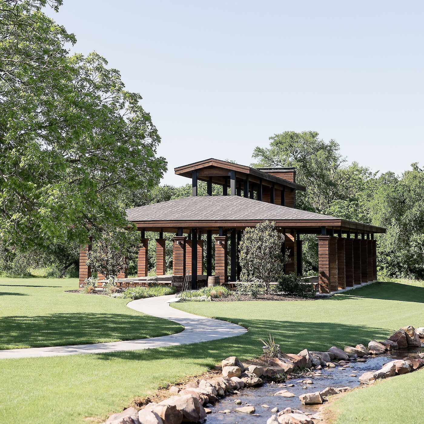 Modern outdoor wooden pavilion or gazebo wedding structure with a layered roof, set in a sunny, manicured green park. A winding path leads to the structure, passing a small stream lined with rocks.