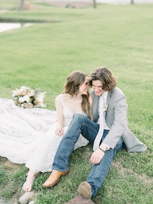 Sweet wedding portrait of the couple seated barefoot on a grassy bank near a pond. The groom wears a gray jacket and denim jeans with cowboy boots, and the bride wears a sheer lace gown, looking affectionately at the groom.