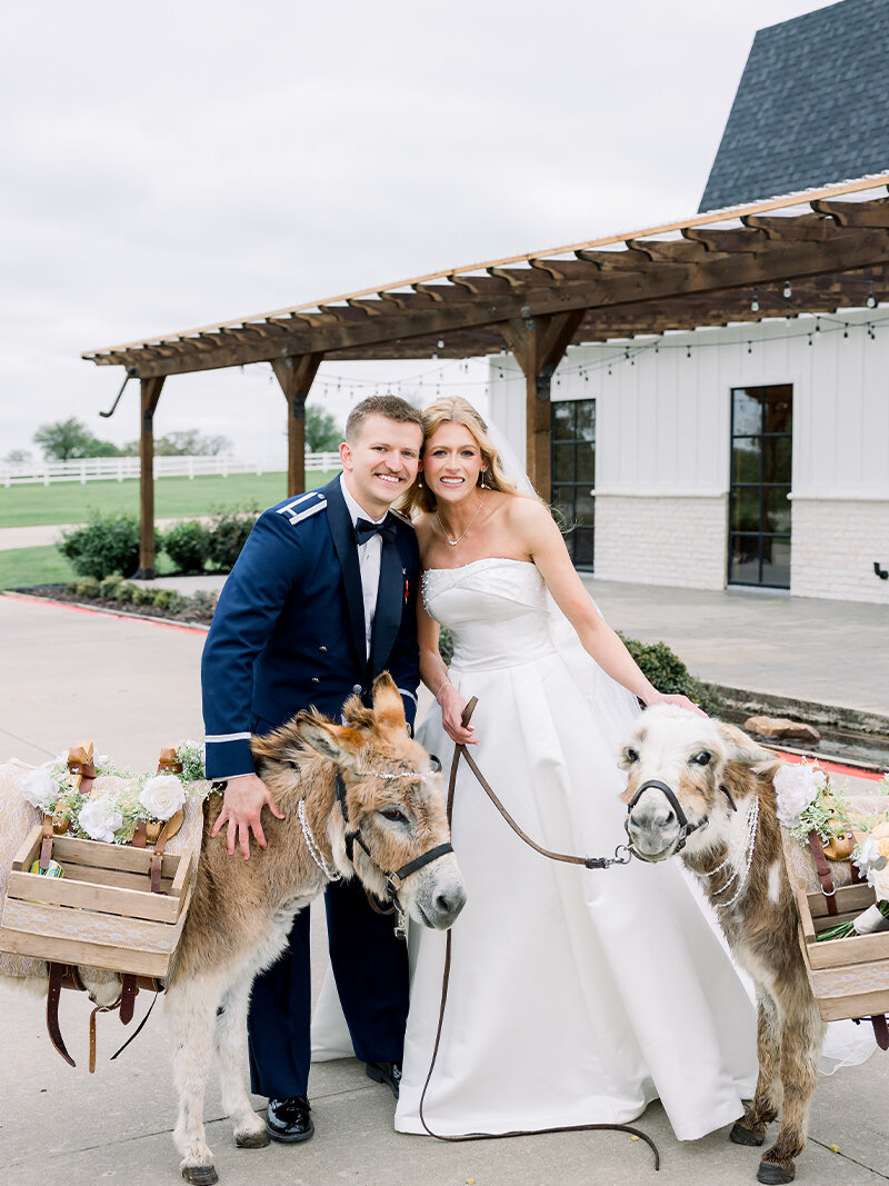 Black and white photo of the bride and groom kissing next to their tiered wedding cake. Both are holding champagne flutes, standing against a rustic wood-plank wall in a barn venue.