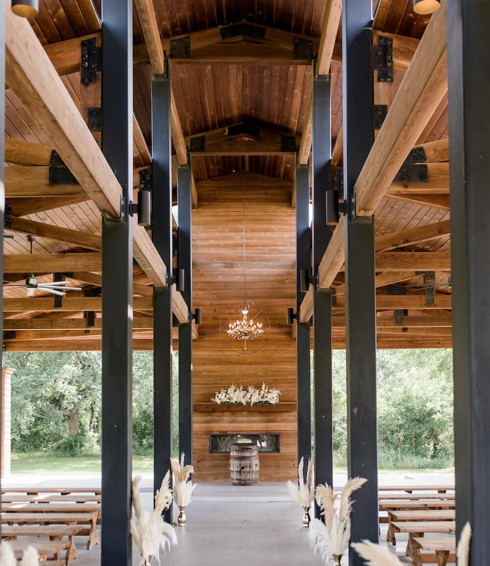 An open-air wedding ceremony pavilion with a wood ceiling, dark support beams, rows of wooden benches, and pampas grass lining the aisle.