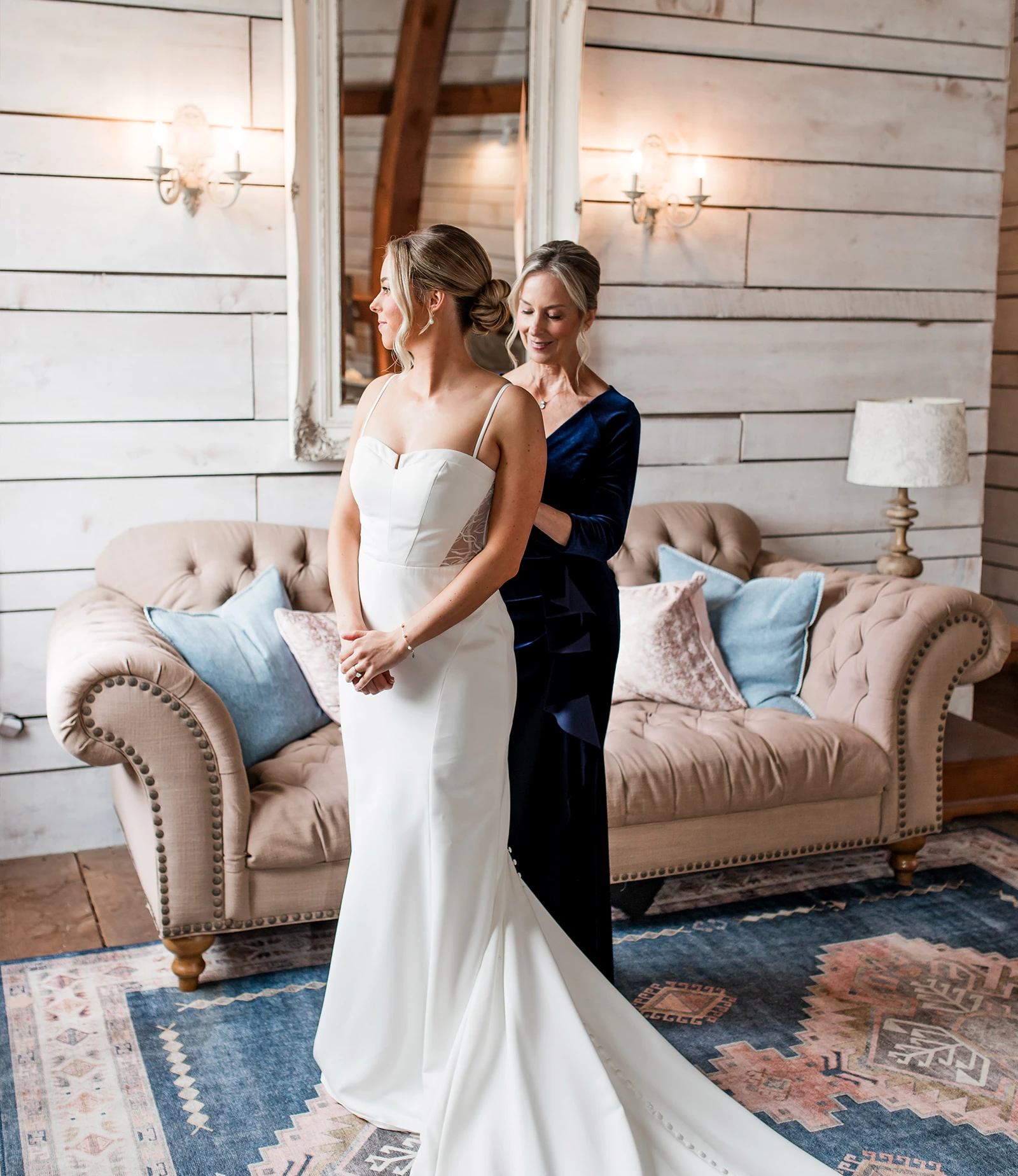 Warm and rustic wedding reception setup inside a barn venue with knotty pine walls and a ceiling featuring wood beams. Round tables are set with white linens, pink runners, and brown folding chairs, with string lights draped around wooden pillars.