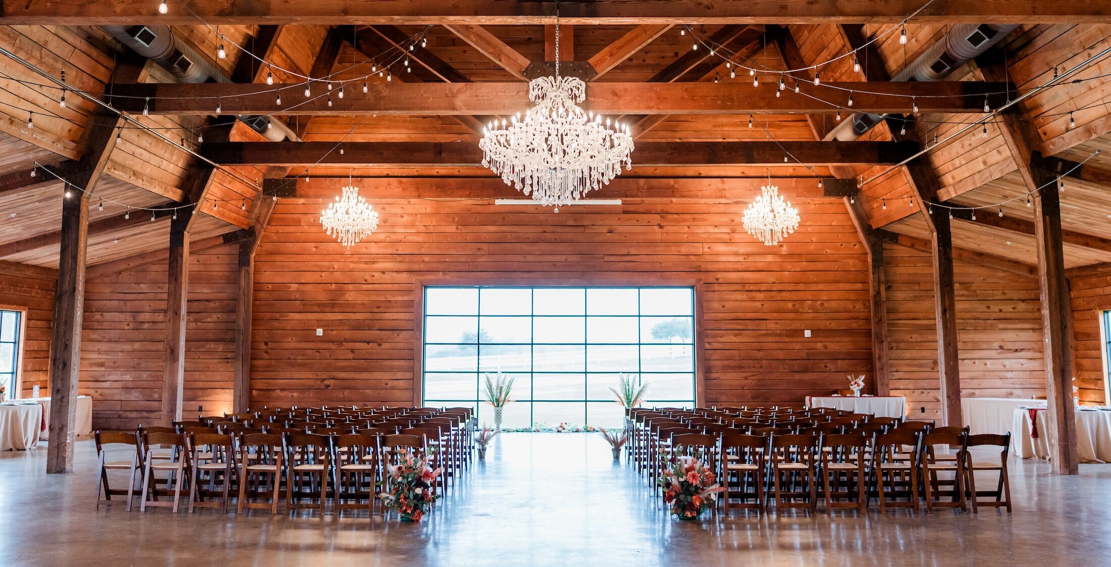 Grand rustic wedding ceremony setup inside a barn venue with high vaulted ceilings, exposed wooden beams, and horizontal wood-plank walls. The aisle is lined with dark wooden chairs and bistro lights lead to an altar with a low horizontal window, lit by crystal chandeliers.