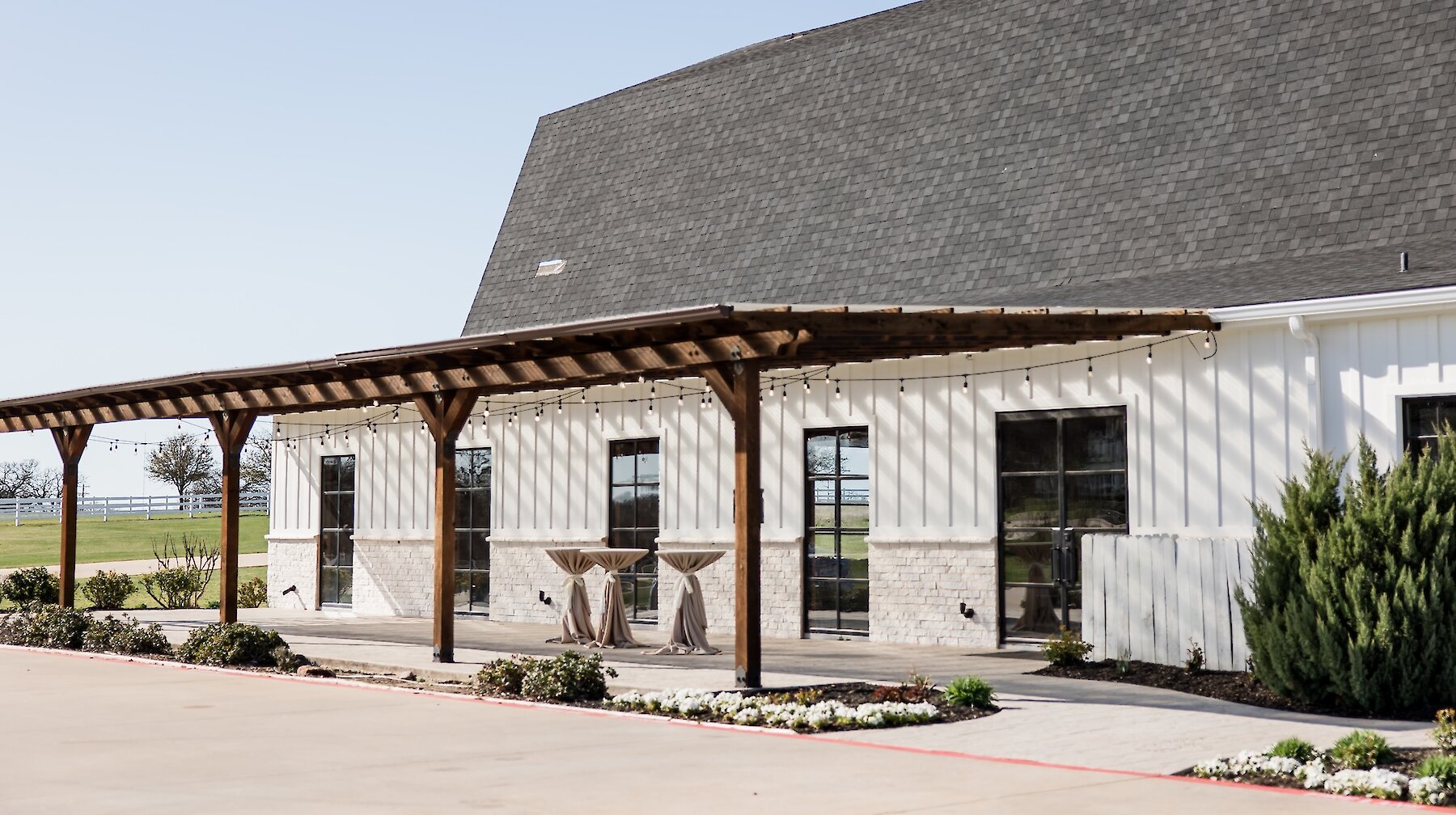 The modern exterior of a white barn venue featuring a wooden pergola-covered patio with cocktail tables and string lights.