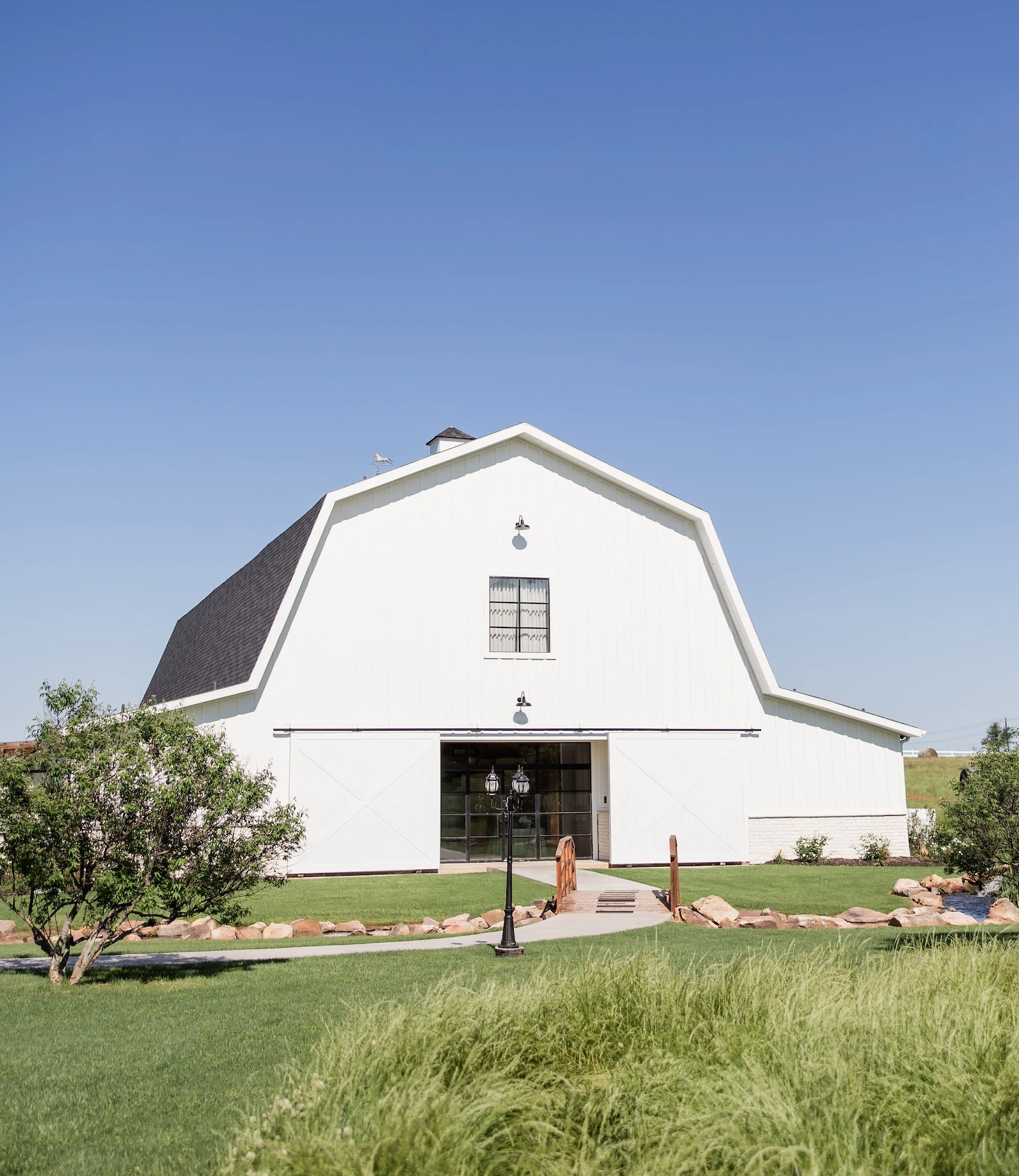 White modern barn wedding venue with sliding doors, manicured green lawn, stone-lined walkway, and clear blue sky