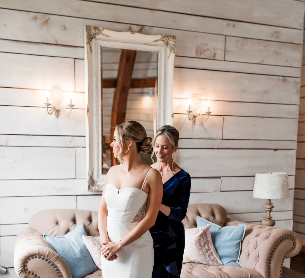 The bride, wearing a white spaghetti strap gown, is being helped into her dress by a bridesmaid in a velvet blue dress in a rustic bridal suite.