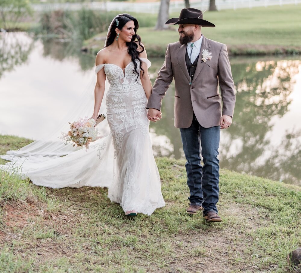 Romantic Western-style wedding portrait of the couple holding hands and walking by a pond at sunset. The groom wears a cowboy hat and brown jacket with denim jeans, and the bride wears an off-the-shoulder illusion lace gown.
