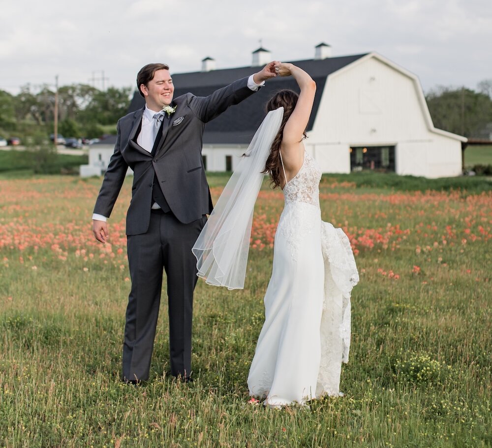 Joyful wedding portrait of the groom spinning the bride in a wildflower field with red-orange blooms. The groom wears a dark gray suit, and the background features a white barn venue under a cloudy sky.