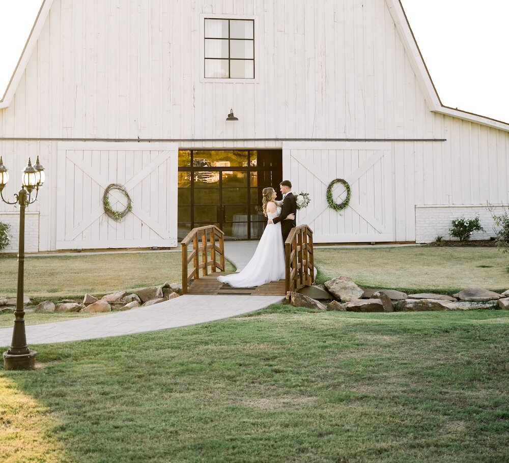 Bride and groom embracing on a small wooden bridge in front of a large white barn wedding venue at sunset