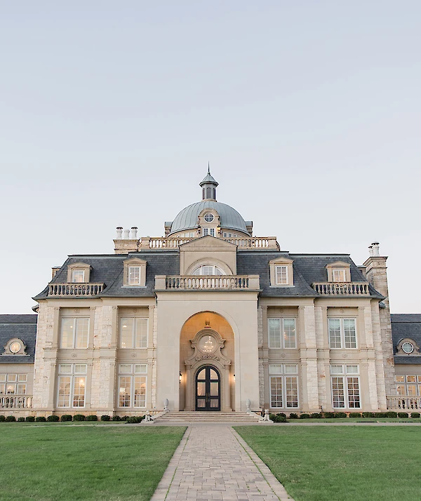 Grand European-style mansion with a central dome, arched entrance, slate roof, and manicured landscaping with rounded hedges and trees on a sunny day.