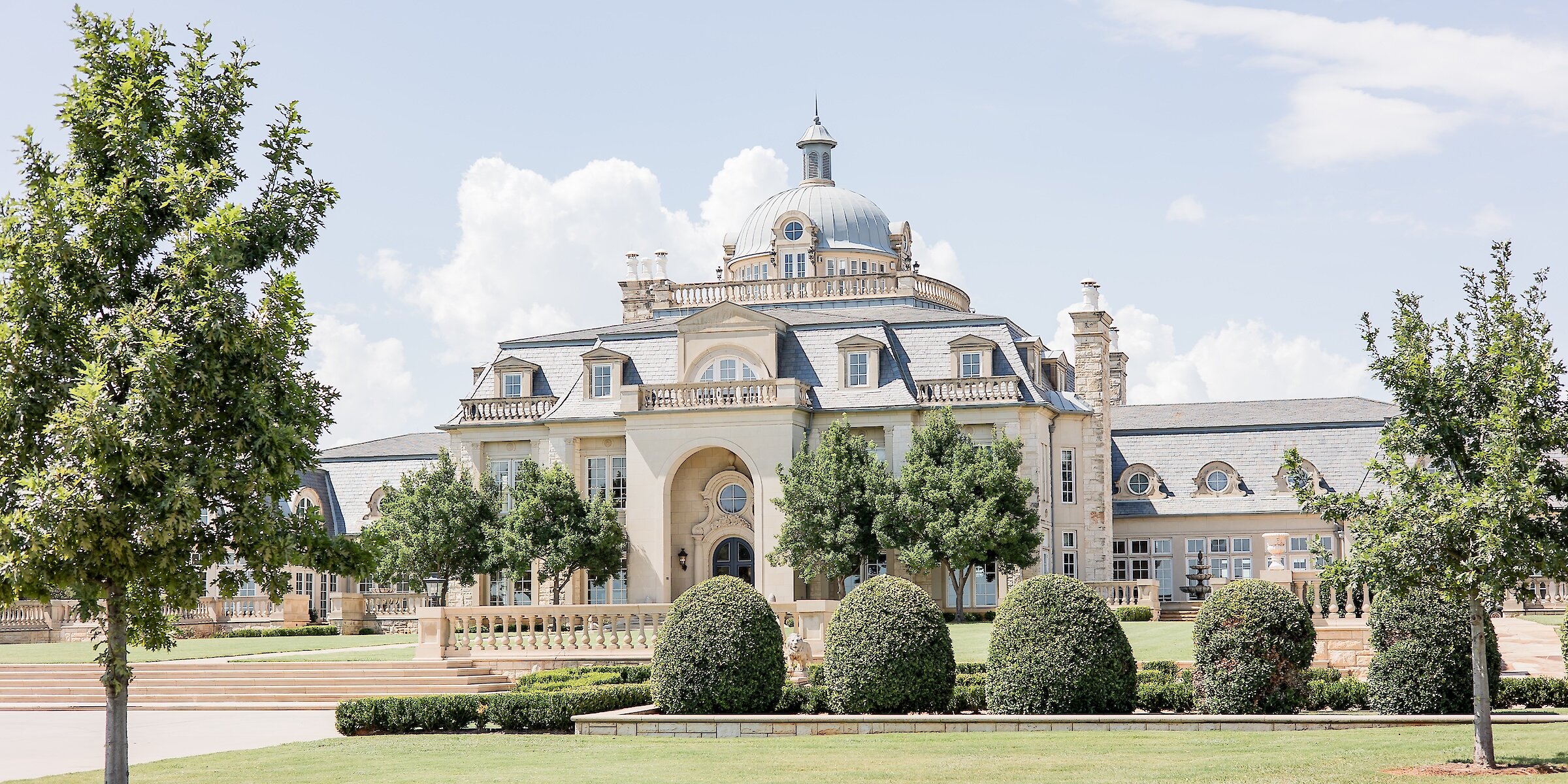 Grand European-style mansion with a central dome, arched entrance, slate roof, and manicured landscaping with rounded hedges and trees on a sunny day.