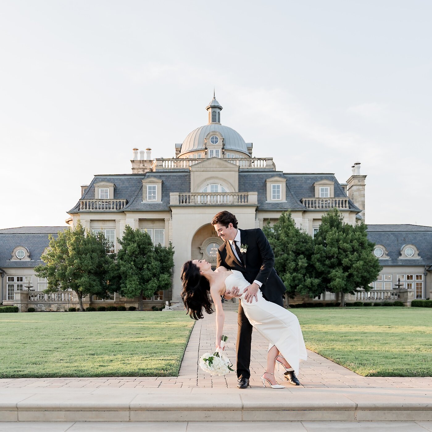 Groom dipping the bride in front of a grand estate at sunset as she holds a bouquet and smiles up at him