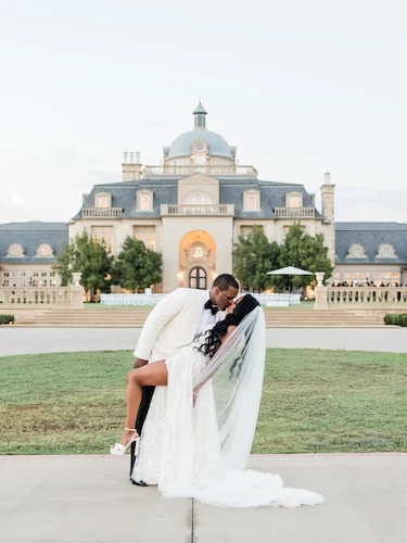 Groom dipping the bride for a kiss in front of a grand French-chateau style wedding venue at sunset.