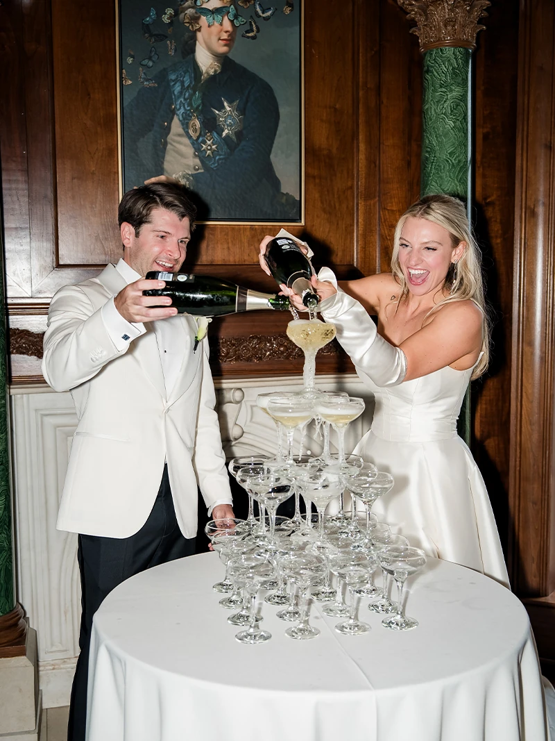 Bride and groom sharing a kiss in an elegant hallway with large windows and a dramatic ruffled wedding gown trailing behind.
