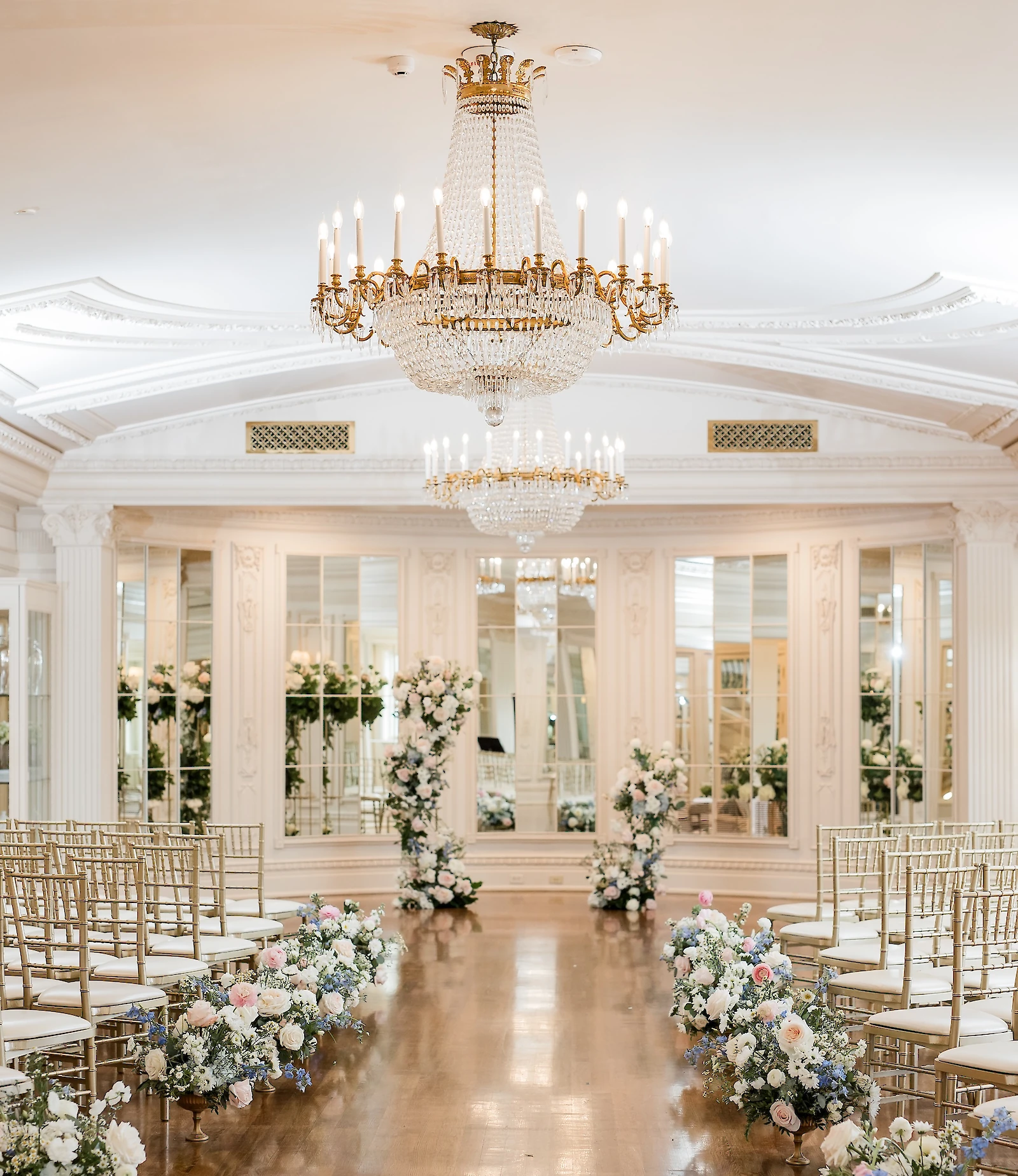 An elegant indoor ceremony space with gold chiavari chairs, crystal chandeliers, mirrored walls, and pastel floral arrangements lining the dark wood aisle.