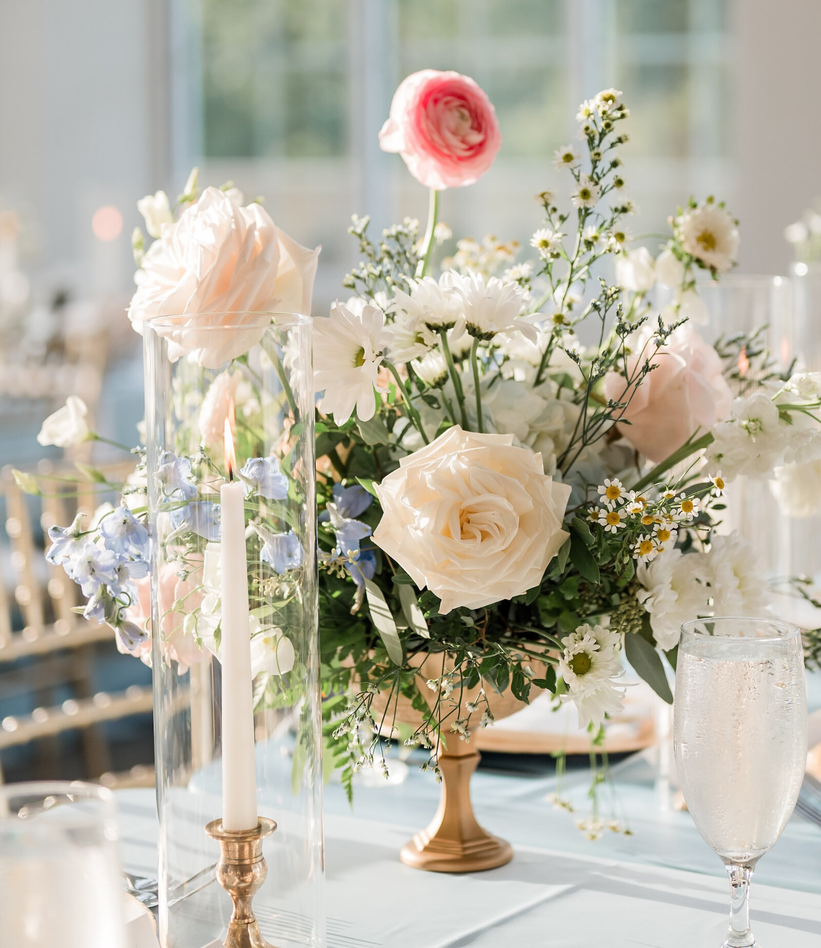 Elegant wedding centerpiece with pastel roses, daisies, and greenery in a gold vase, surrounded by glowing taper candles and glassware in soft natural light.