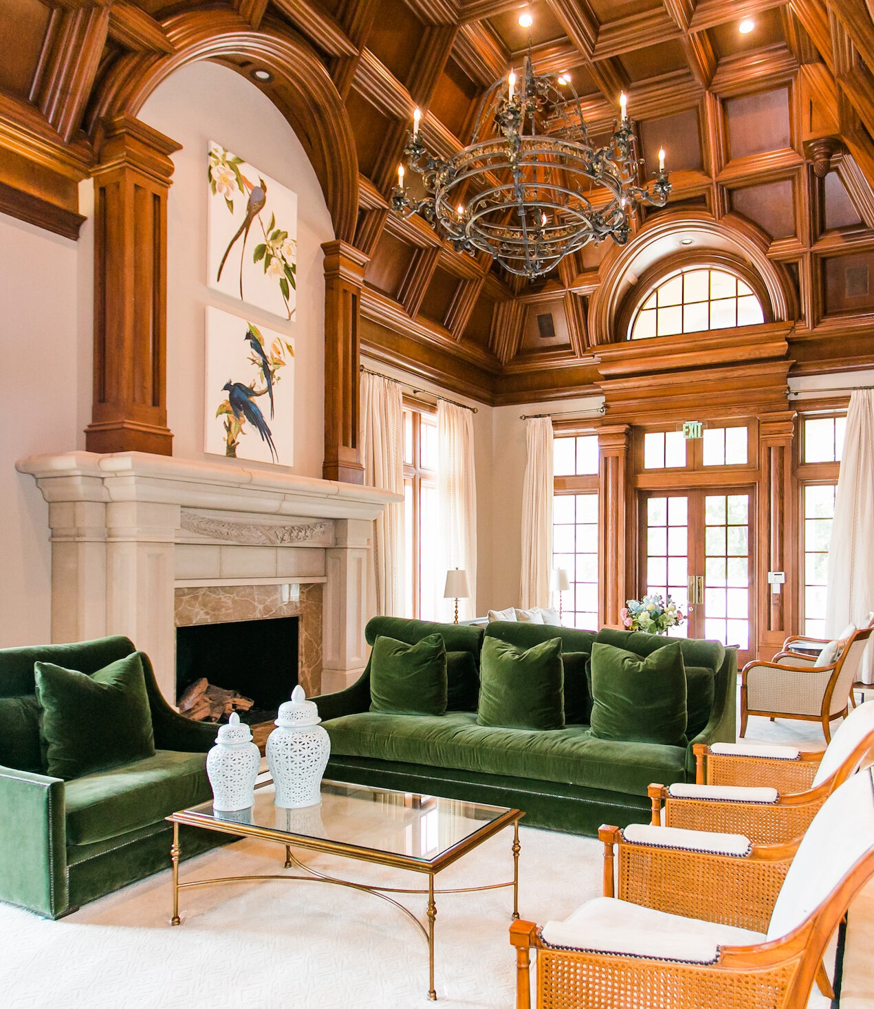 Luxurious living room with coffered wood ceiling, green velvet sofas, ornate chandelier, marble fireplace, and elegant French doors.