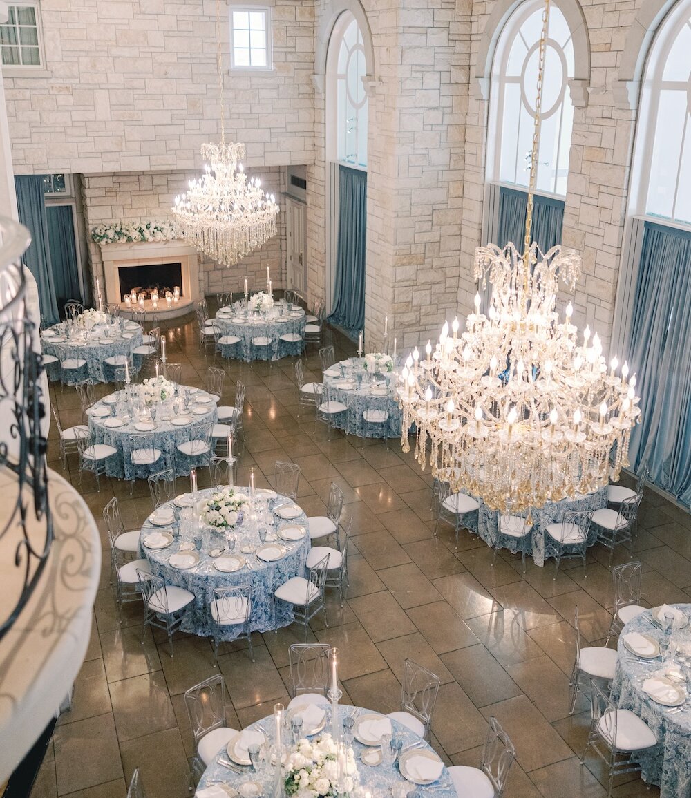 An aerial view of a luxury stone ballroom set for a reception with blue patterned tablecloths, acrylic chairs, and large crystal chandeliers.