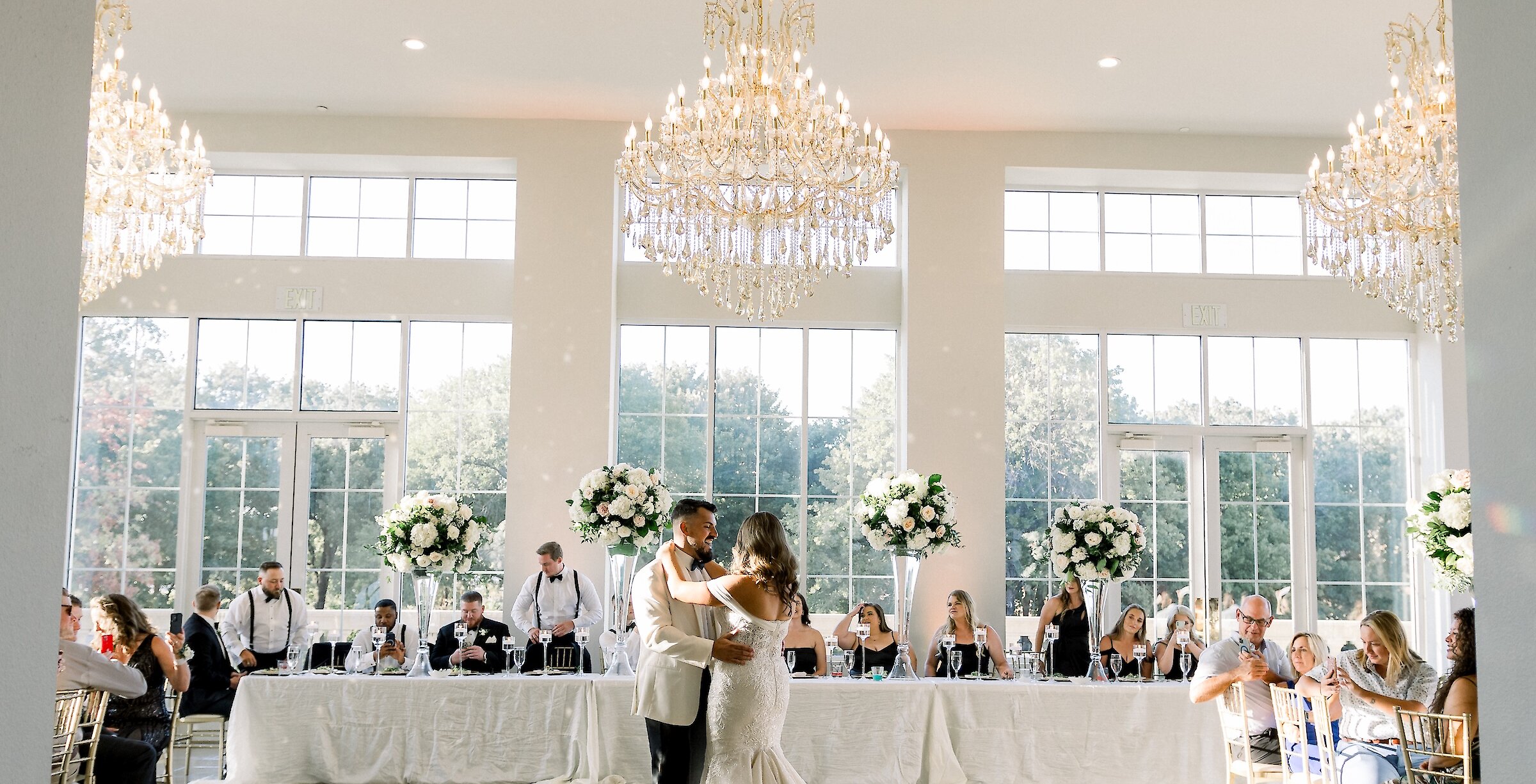 The bride and groom share their first dance in a large, brightly lit ballroom with tall windows, crystal chandeliers, and a long head table.