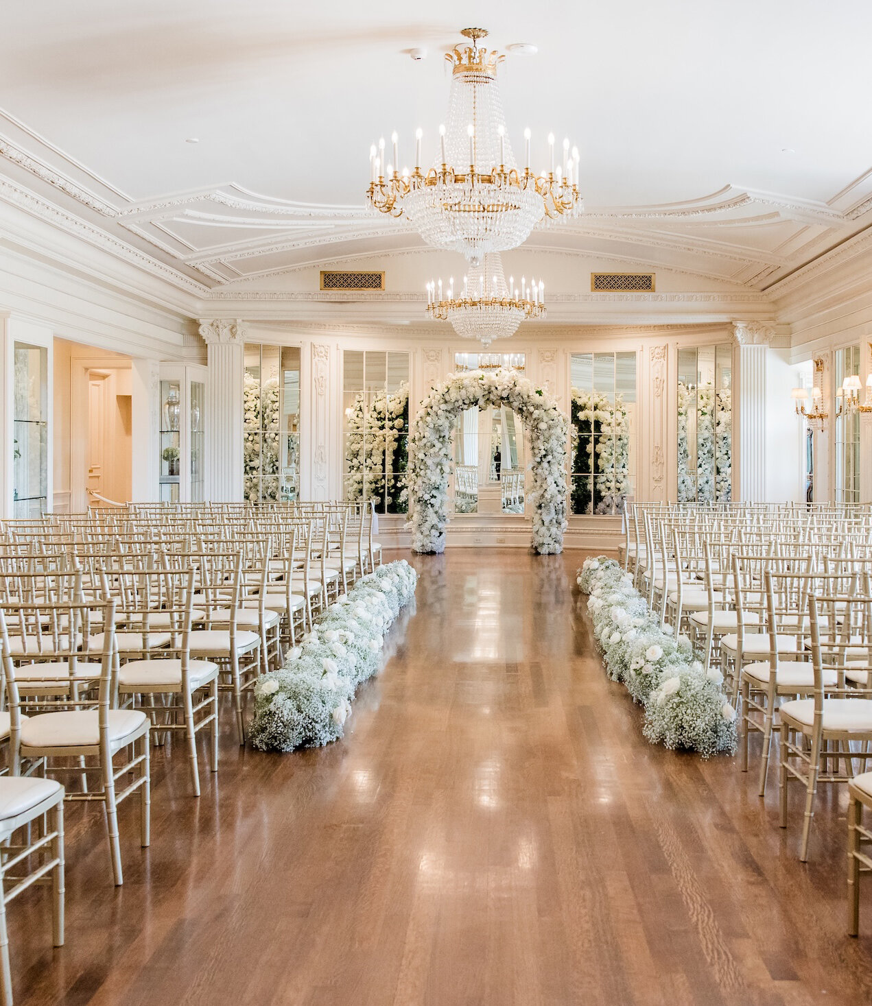 Luxurious indoor wedding ceremony setup with gold chairs, white floral aisle arrangements, and a grand white flower arch beneath ornate chandeliers in an elegant ballroom