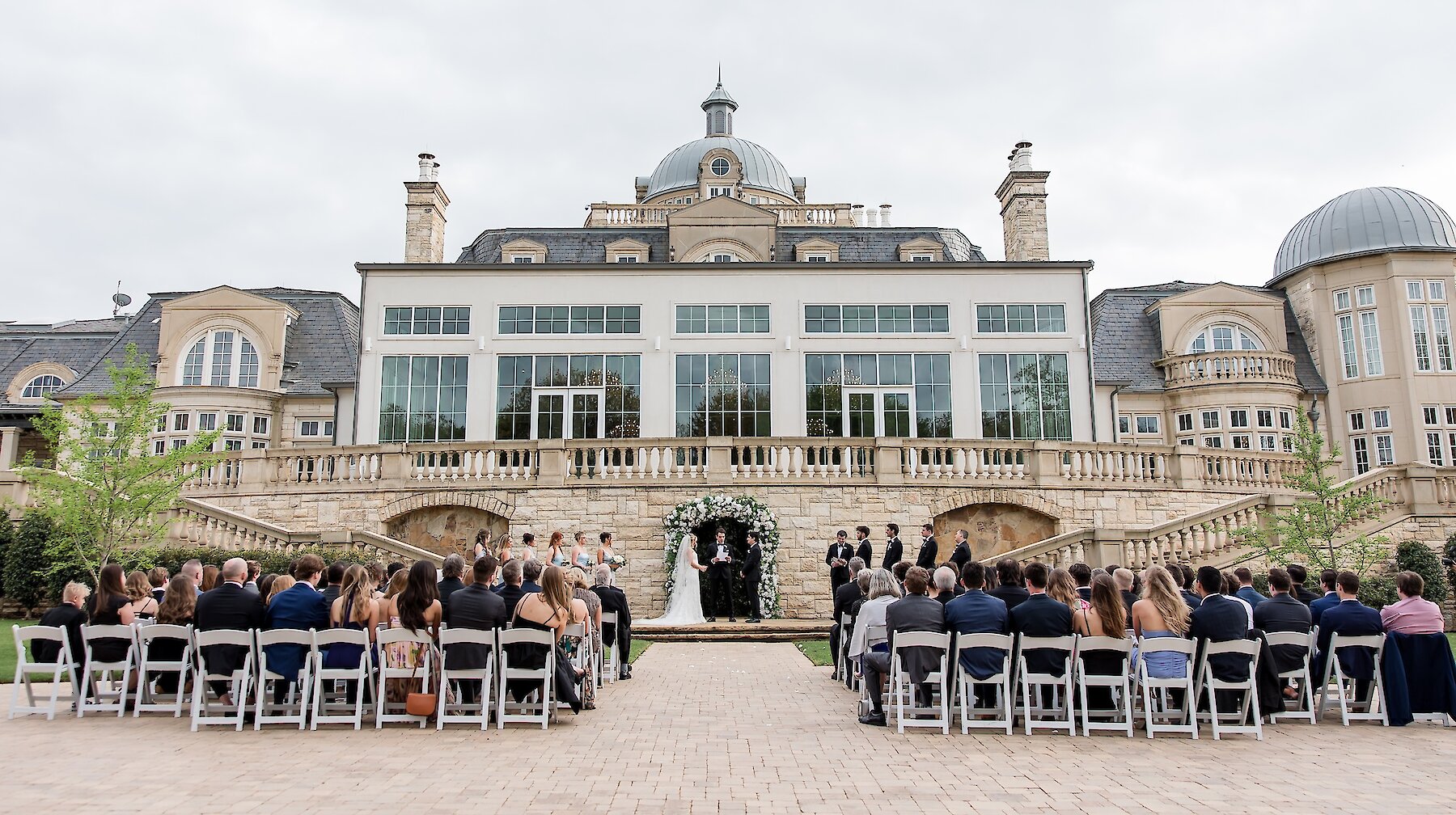 outdoor wedding ceremony at a grand estate with the couple standing beneath a floral arch and guests seated facing the mansion