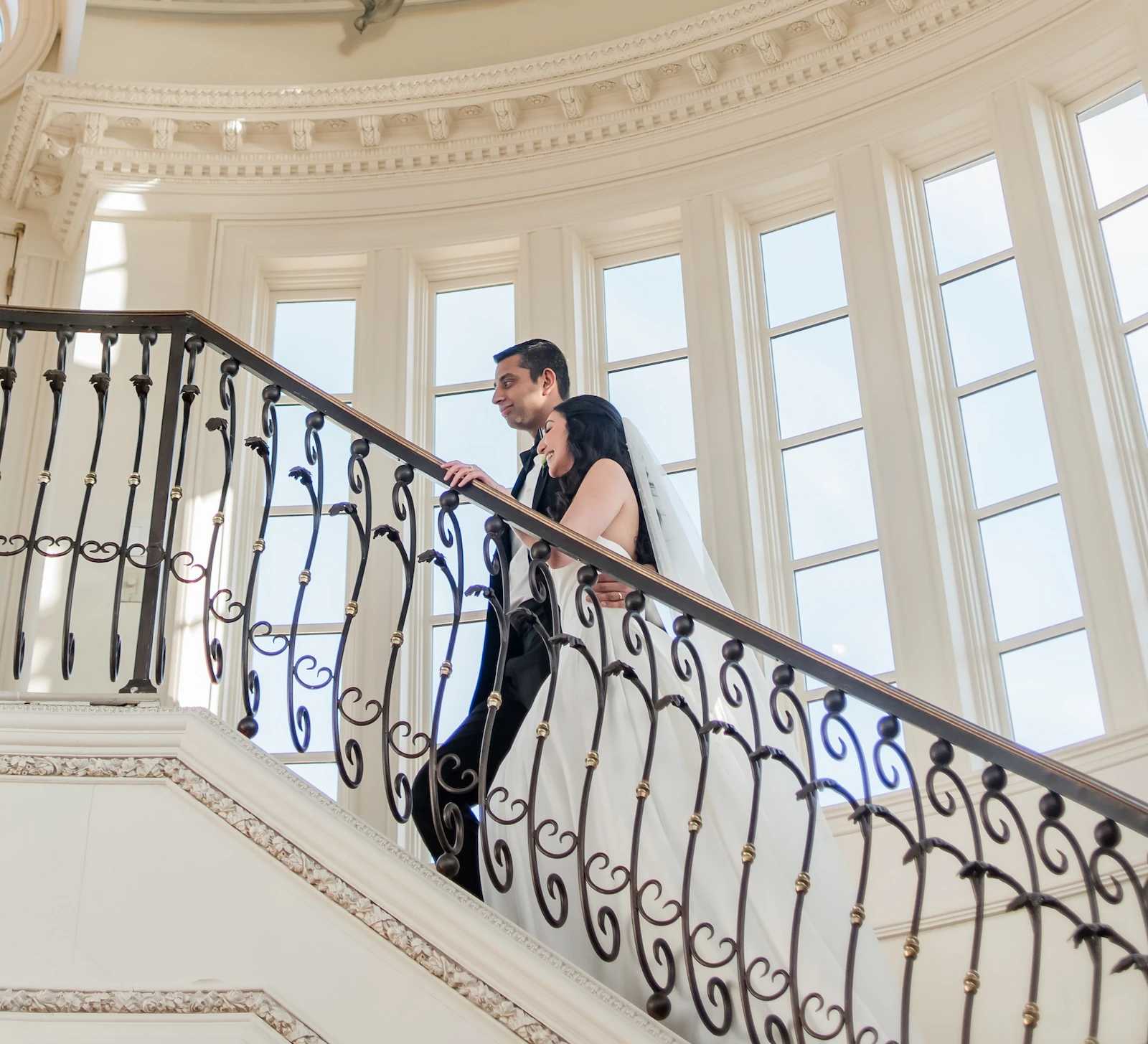 The bride and groom pose on a grand winding staircase with a wrought iron railing, under a beautiful painted dome ceiling mural of a sky.