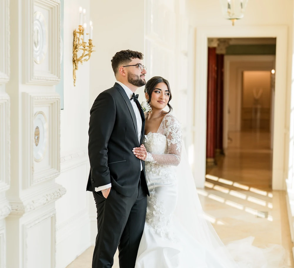 Bride and groom standing together in an elegant hallway with ornate white detailing, soft natural light, and the bride in a long-sleeve lace gown.