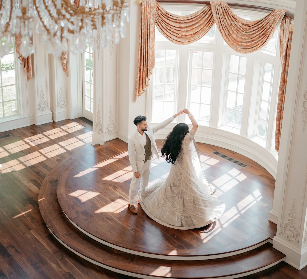 An overhead shot of a bride and groom dancing on a round, tiered dark wood floor in a grand room with tall windows and a large crystal chandelier.