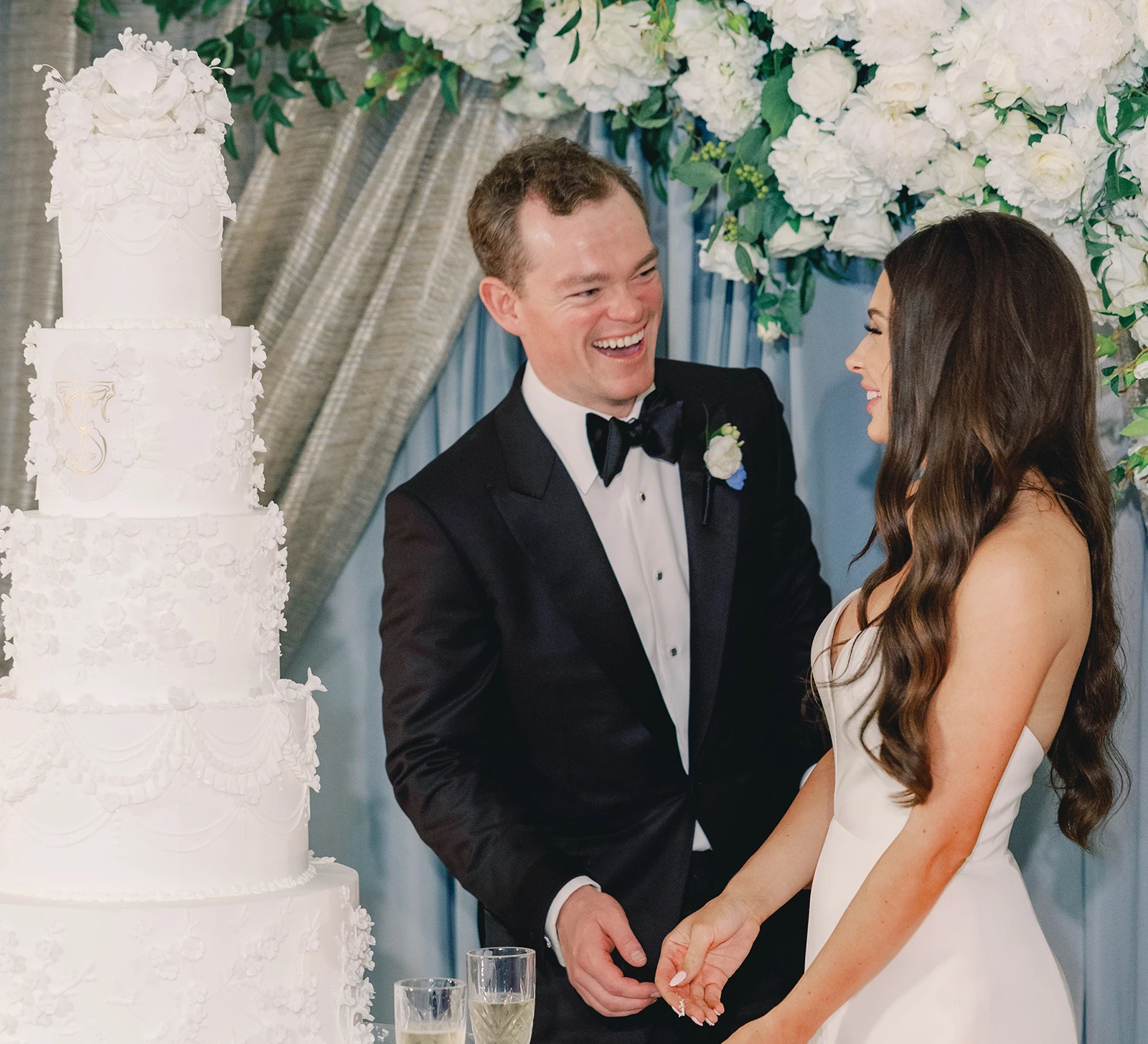 Bride and groom laughing as they cut a tall white wedding cake decorated with intricate floral details, standing beneath a white flower arch